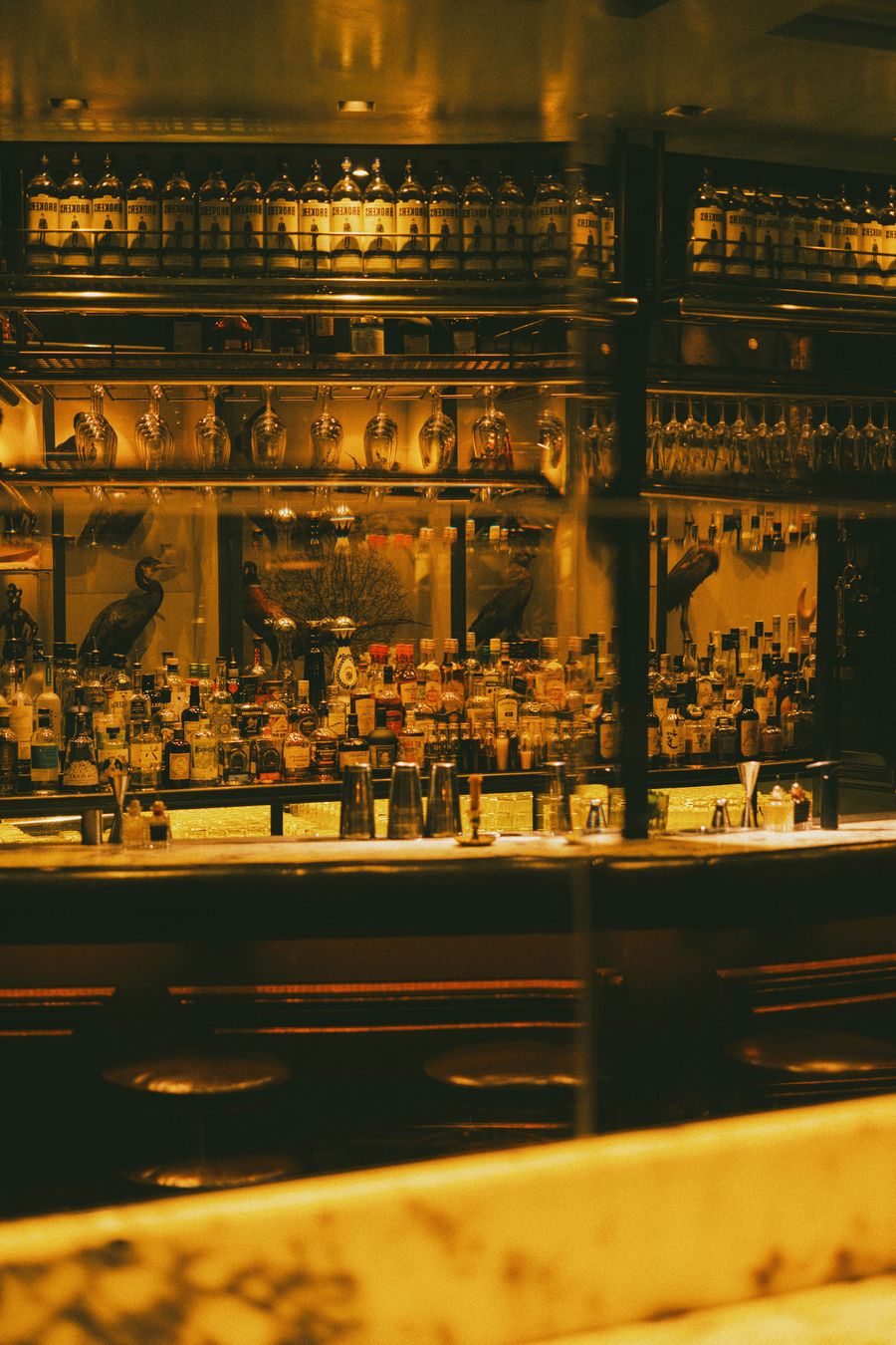 Moody interior of Kyrios cocktail bar in Athens, where amber lighting illuminates densely stocked liquor shelves, rows of hanging stemware and a long, dark bar counter. Mirrored surfaces multiply the bottles and reflections, while decorative bird figures and low leather stools lend the space a richly layered, vintage-inspired character.