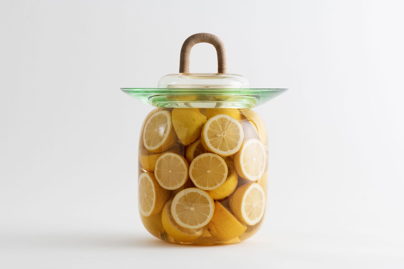 A large clear glass jar filled with lemon halves, closed with a green-tinted glass rim and a clear lid featuring a wrapped handle.