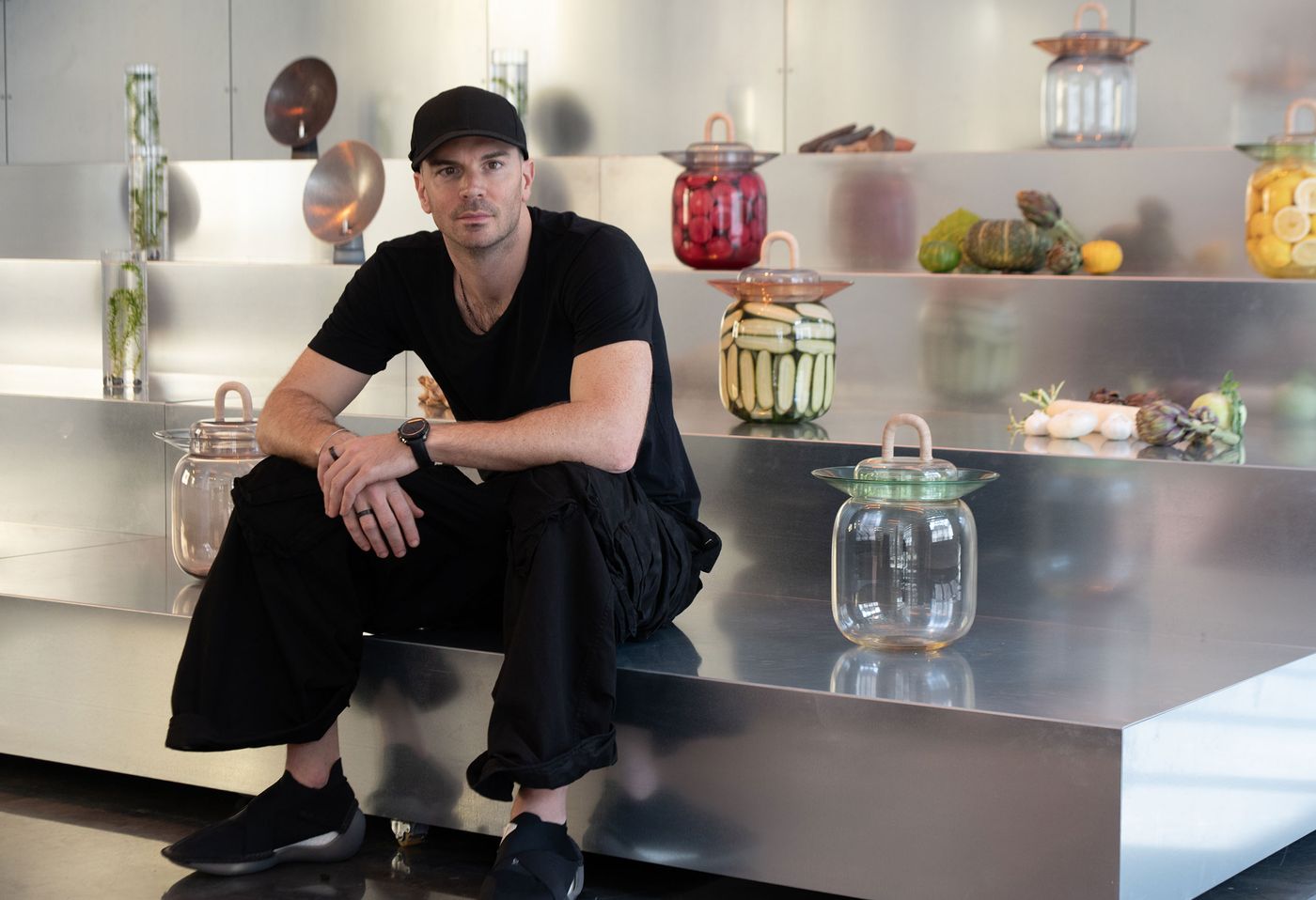Benjamin Hubert dressed in black and sits on a sleek metallic platform surrounded by sculptural glass jars filled with colorful pickled vegetables, showcasing a futuristic yet organic display.