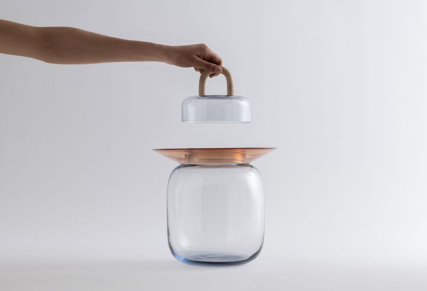 A hand lifts a delicate glass lid with a wrapped handle above a sculptural glass vessel with a wide amber-hued rim, against a soft white background.