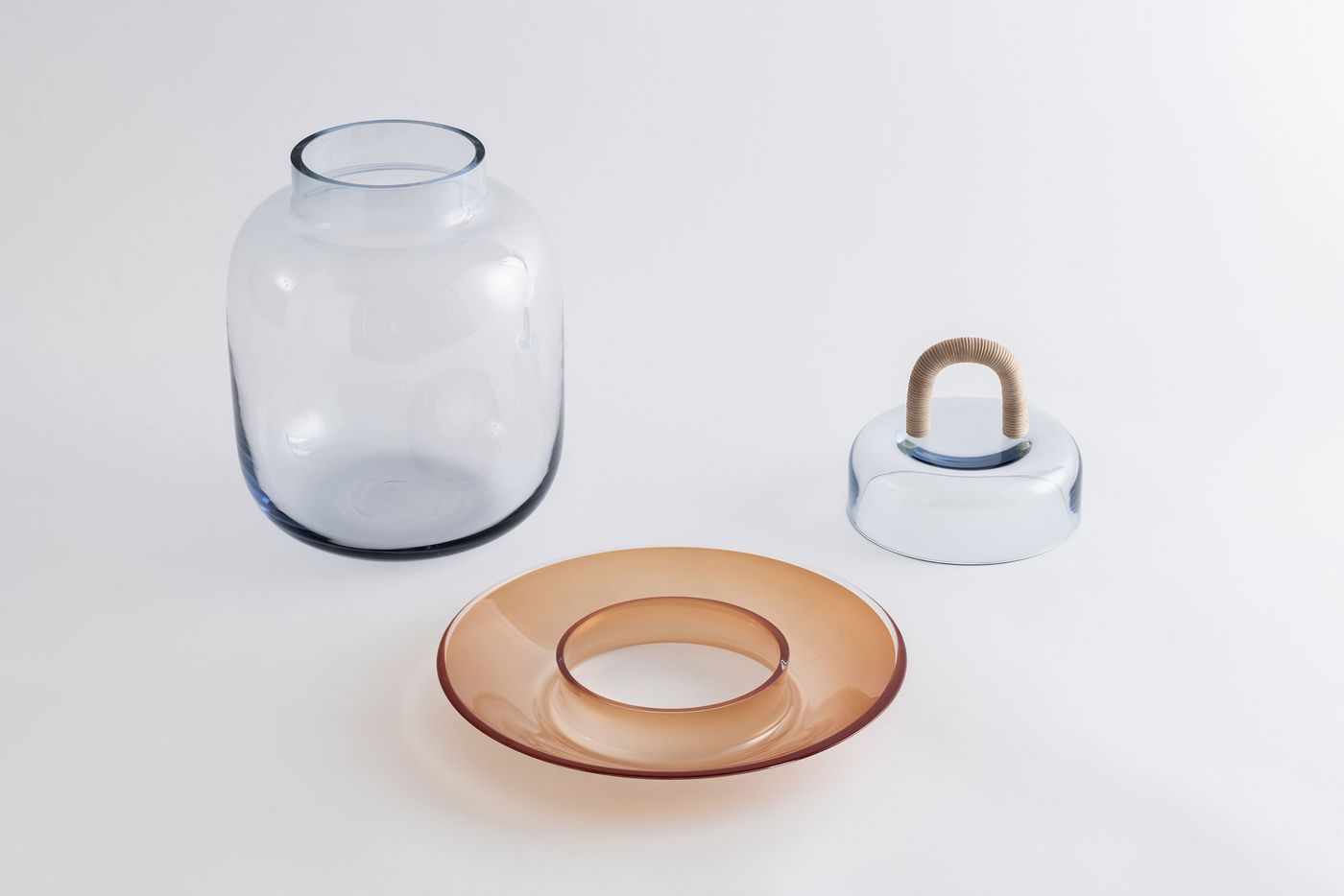 Three separate pieces of the glass vessel — a clear jar, an amber disk, and a clear lid with a wrapped handle — laid out elegantly on a white surface.
