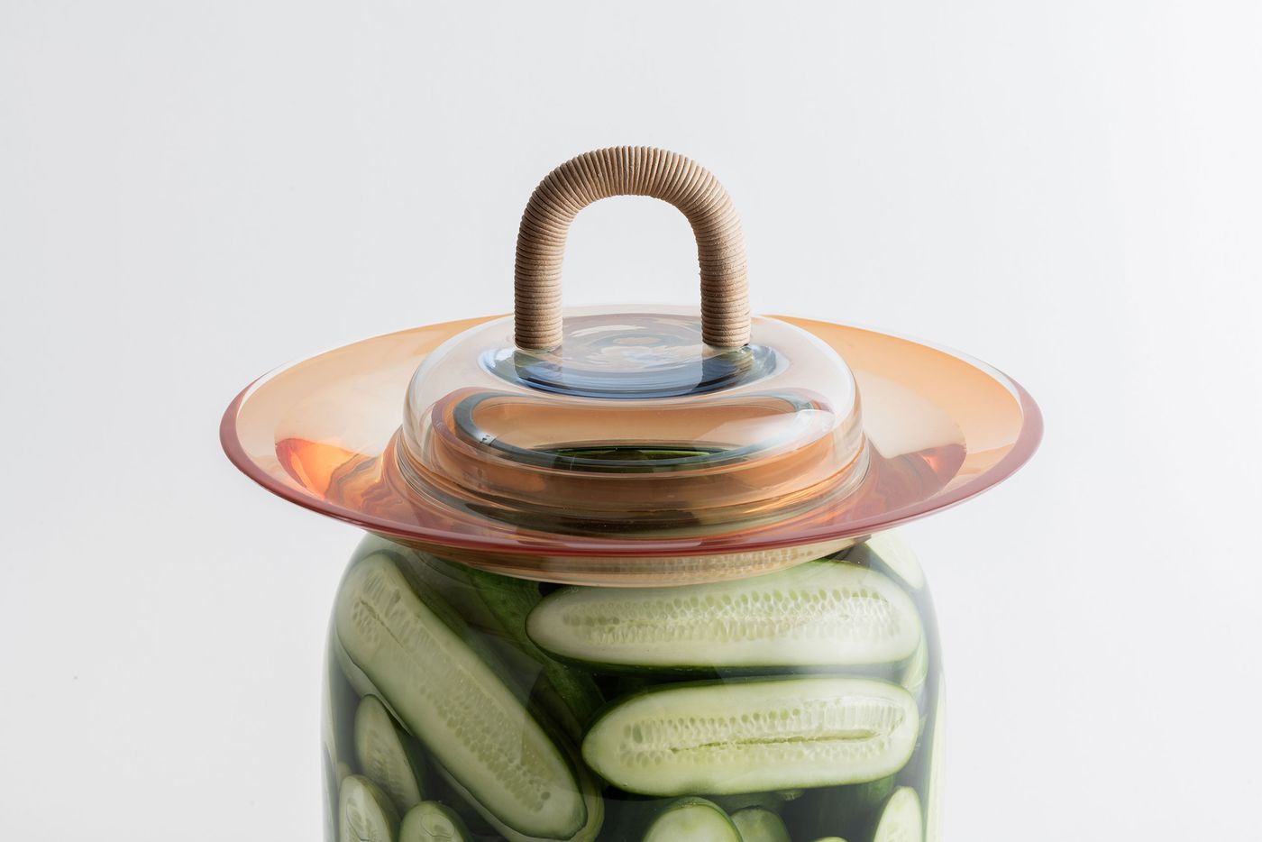 A clear glass jar filled with pickled cucumbers, topped with an amber-hued rim and a transparent lid with a tactile wrapped handle.