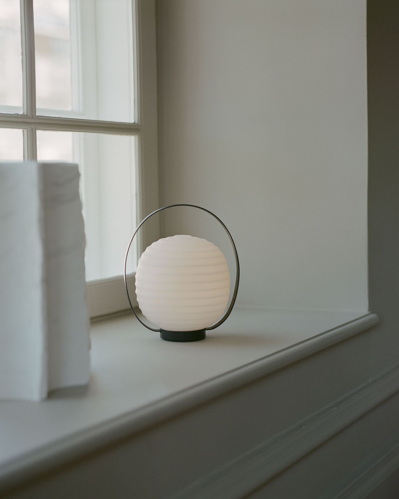 A minimalist white spherical lantern with horizontal ridges, encased in a thin black metal handle, placed on a light-colored windowsill next to two stacked white books, with soft natural light coming through the window.