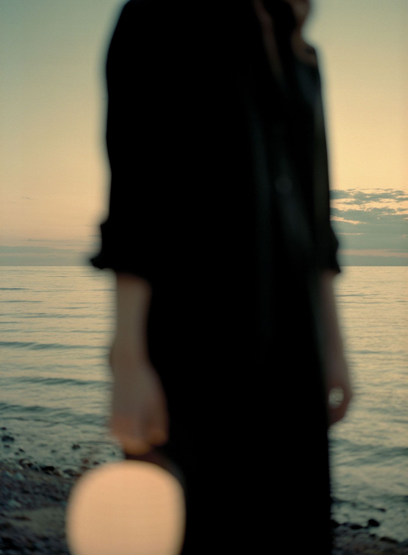 A blurred image of a person dressed in black standing near a rocky shoreline at sunset, holding a glowing spherical lantern with a black handle close to the camera, with the calm sea and a cloudy sky in the background.