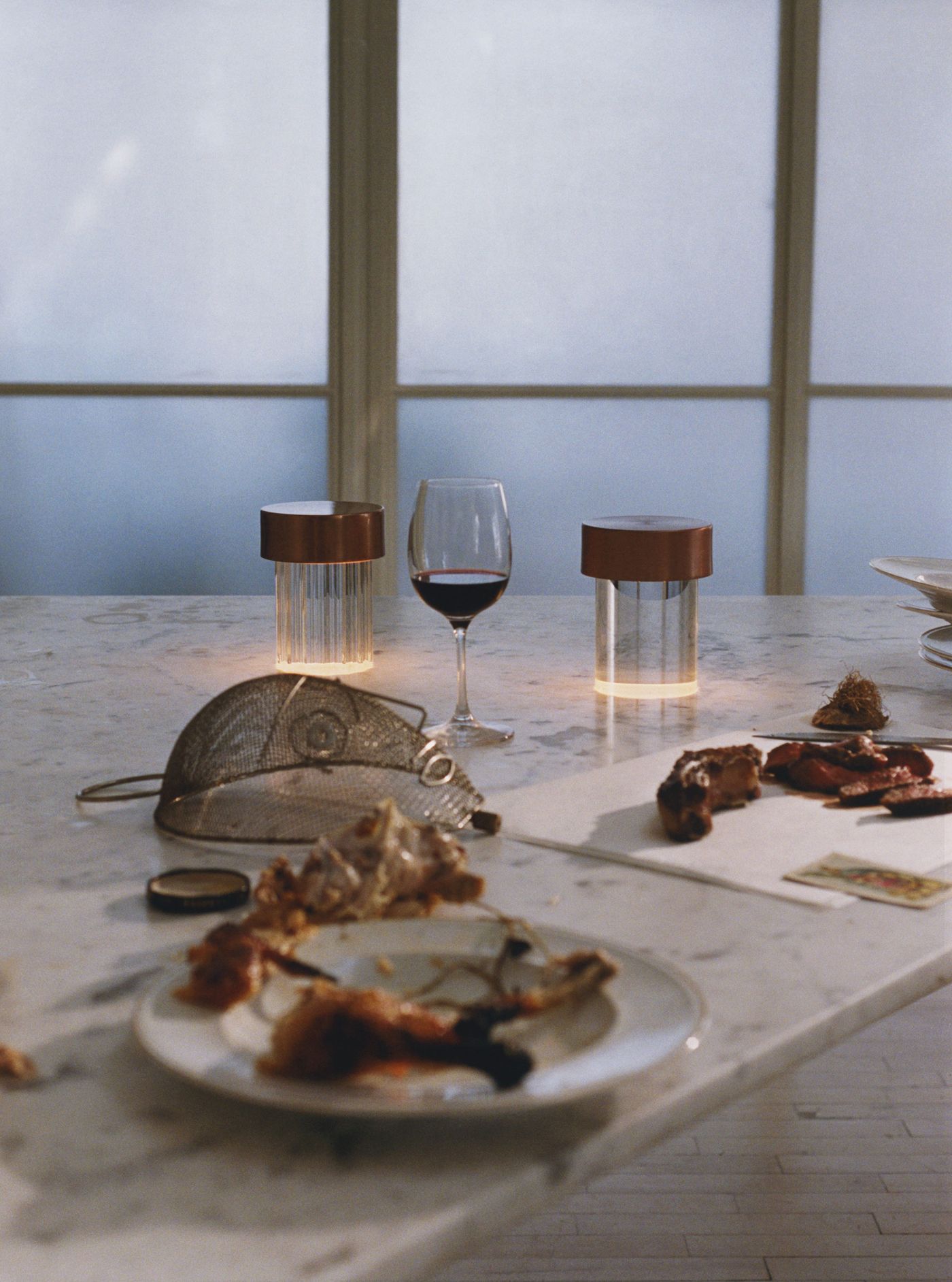 Two copper-topped Last Order lamps glow gently on a white marble dining table scattered with remnants of a hearty meal—wine, crab claws, and cutlery frozen in a cinematic still life.