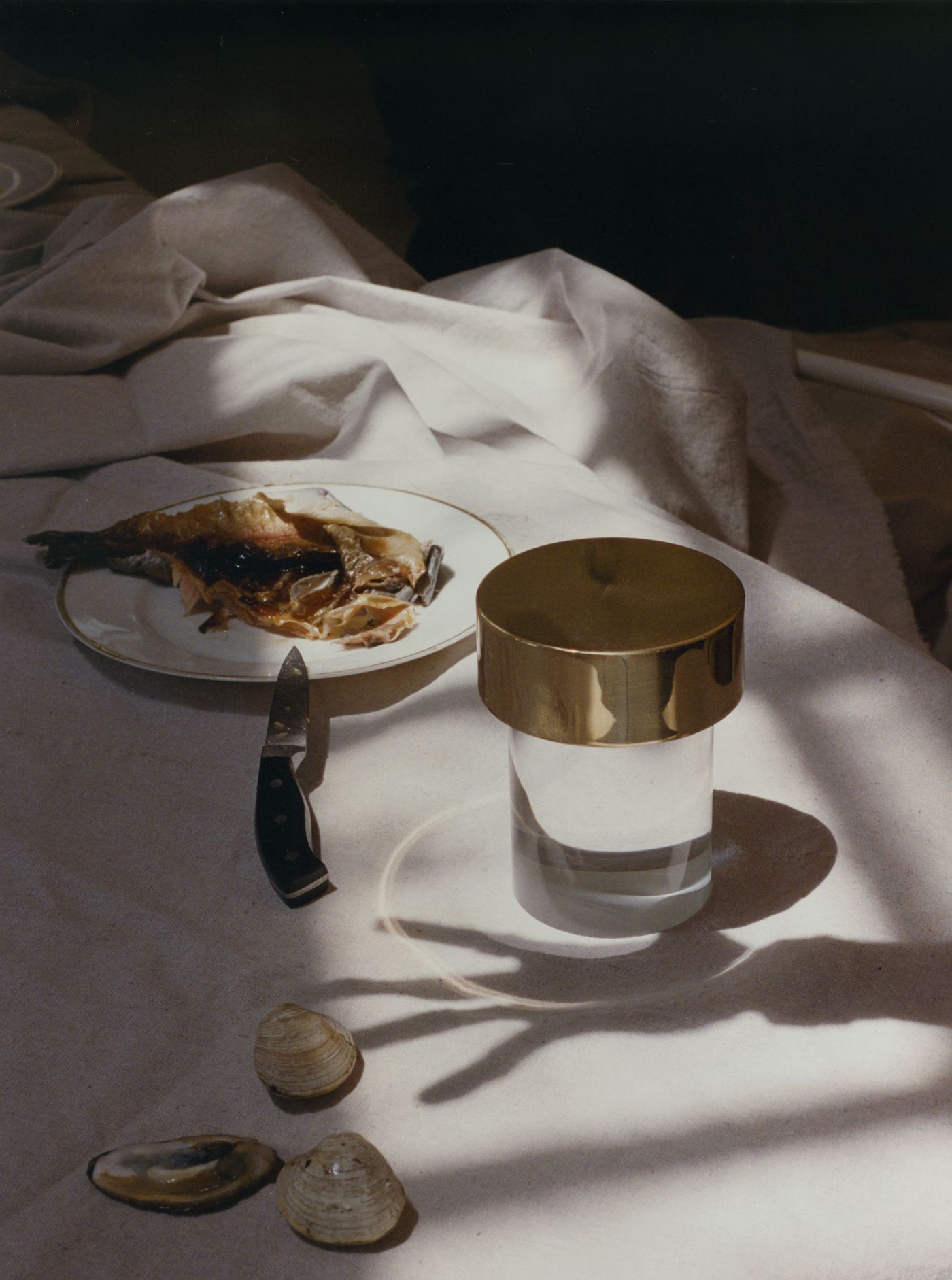 A golden Last Order lamp radiates soft light over a white tablecloth strewn with clamshells, a grilled fish, and a knife—evoking an elegant, rustic seafood feast.