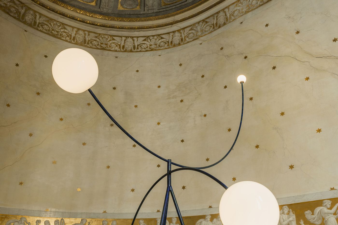 Three spherical white lights float against a grand domed ceiling, adorned with golden stars and classical reliefs, creating a poetic tension between architecture and contemporary lighting. Three spherical white lights float against a grand domed ceiling, adorned with golden stars and classical reliefs, creating a poetic tension between architecture and contemporary lighting.