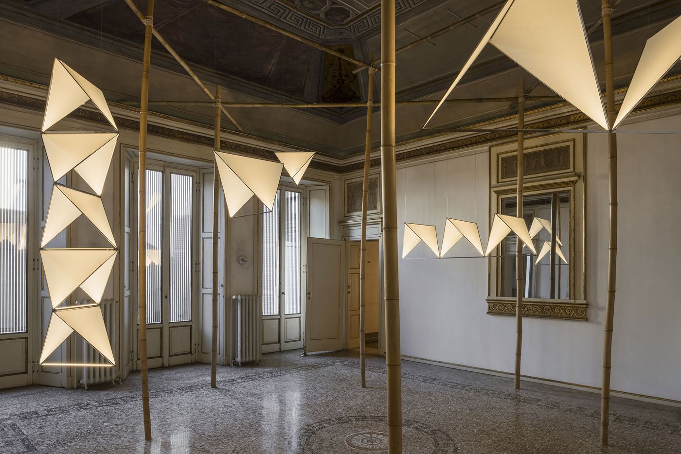 A geometric grid-like light fixture made of polished metal and soft illumination hovers elegantly in a corner with ornate, faded frescoed ceilings. A geometric grid-like light fixture made of polished metal and soft illumination hovers elegantly in a corner with ornate, faded frescoed ceilings.
