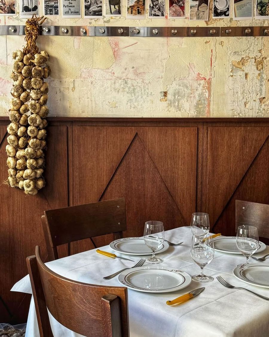 An intimate dining corner at Manári taverna in Athens, featuring dark timber panelling, distressed plaster walls and a long braid of garlic hanging beside a wall of framed photographs. A white tablecloth, traditional crockery, yellow-handled cutlery and sturdy wooden chairs evoke the unpretentious character of an old-school Greek taverna, subtly reinterpreted through contemporary design.