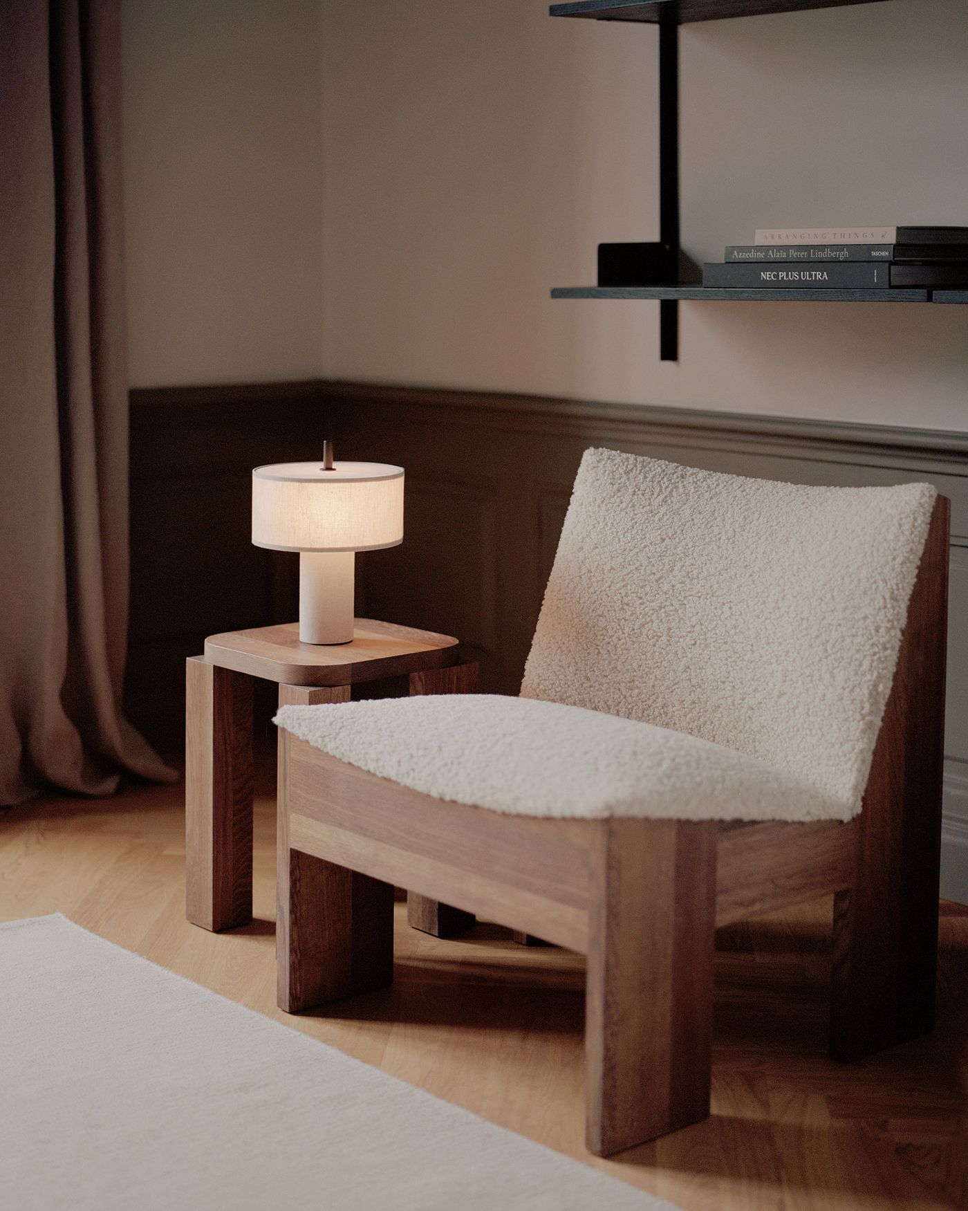 A cozy corner featuring a wooden lounge chair with a thick, textured white cushion and backrest, next to a matching wooden side table holding a modern table lamp with a cylindrical white shade, set against a neutral-toned wall with a floating black shelf displaying a few books.