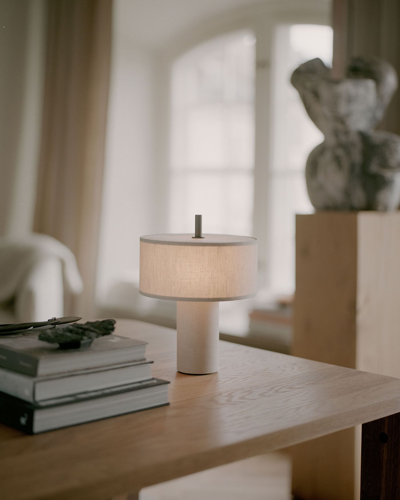 A softly lit, modern table lamp with a cylindrical fabric shade and base sits on a light wooden table next to a stack of books, with a blurred abstract sculpture and large arched window in the background.