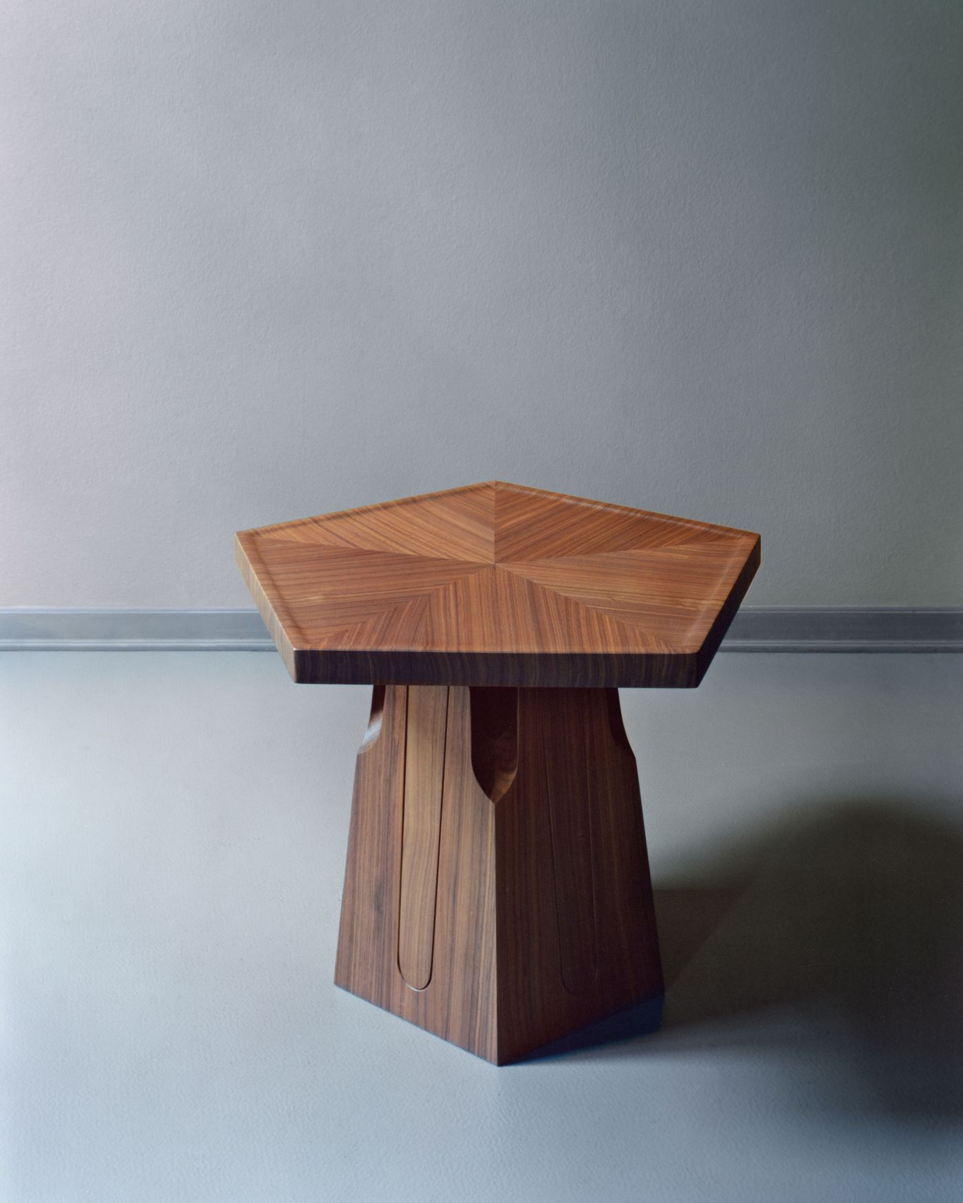 A sculptural wooden side table with a pentagonal top and intricate base detailing, set against a muted grey wall.