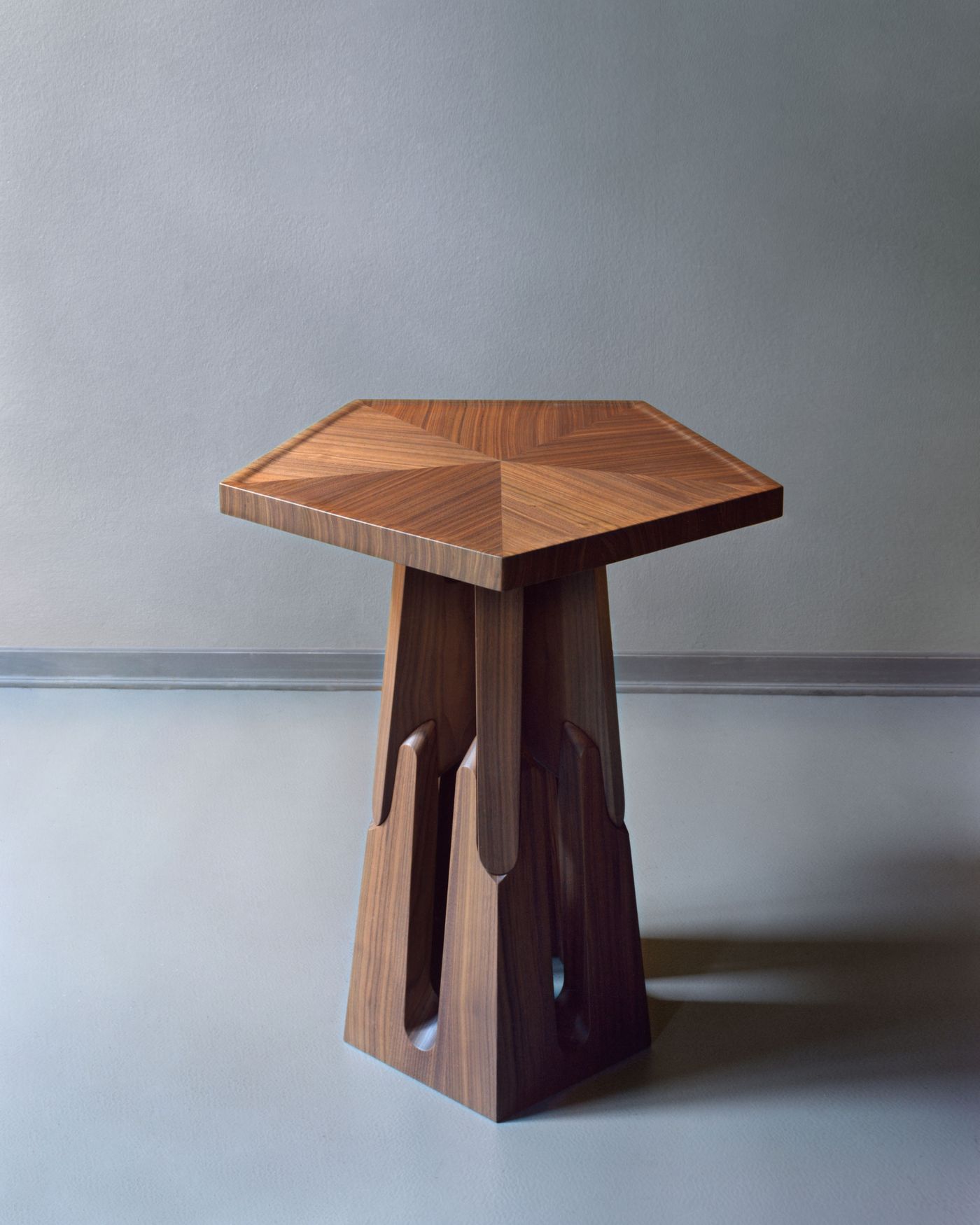 Another sculptural view of the wooden pentagonal table, this time with more dramatic carved base elements against a soft grey wall.