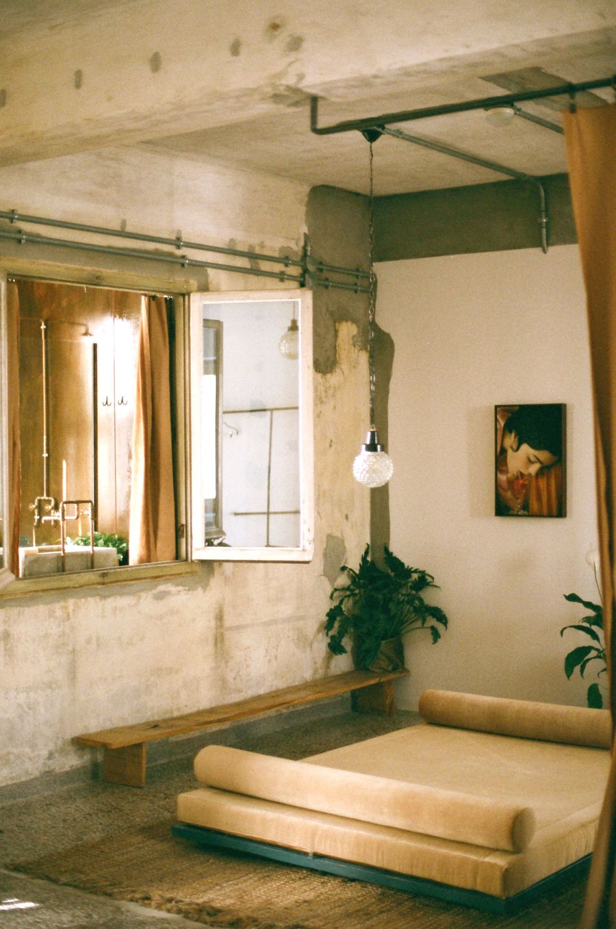 A softly lit interior at Mona boutique hotel in Athens juxtaposes raw concrete surfaces, exposed piping and weathered plaster with a low, cream-coloured daybed and rustic timber bench. An open window, suspended glass pendant and potted greenery lend warmth to the industrial setting, while a small framed portrait adds an intimate, lived-in quality.