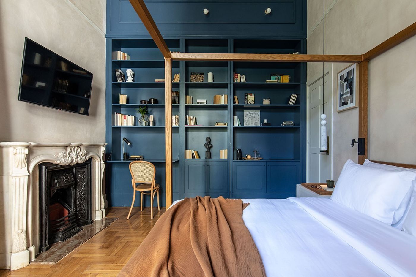 A guest room at Monsieur Didot in Athens combines neoclassical elegance with contemporary comfort. An ornate marble fireplace and parquet flooring retain the townhouse's historic character, while floor-to-ceiling blue bookcases, a rattan chair and a minimalist timber four-poster bed introduce a restrained, residential aesthetic. Books, ceramics and sculptural objects lend the space a cultured, intimate atmosphere.