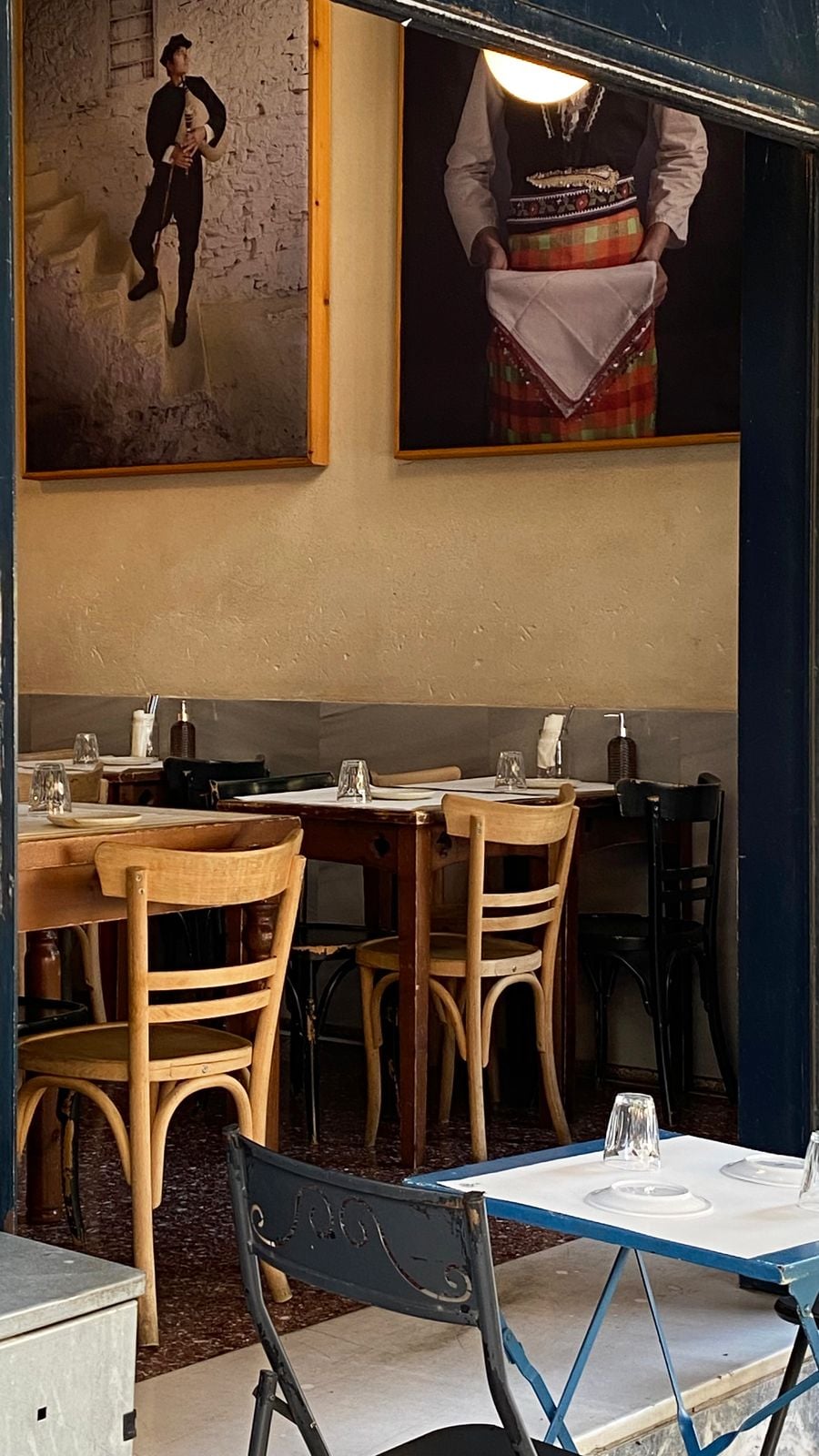 A glimpse inside Mourga restaurant in Thessaloniki reveals an intimate, unpretentious dining room furnished with dark wooden tables and mismatched bentwood chairs. Framed photographs depicting traditional Greek life hang above simply laid tables, while warm lighting and worn surfaces contribute to the restaurant’s nostalgic, lived-in atmosphere.