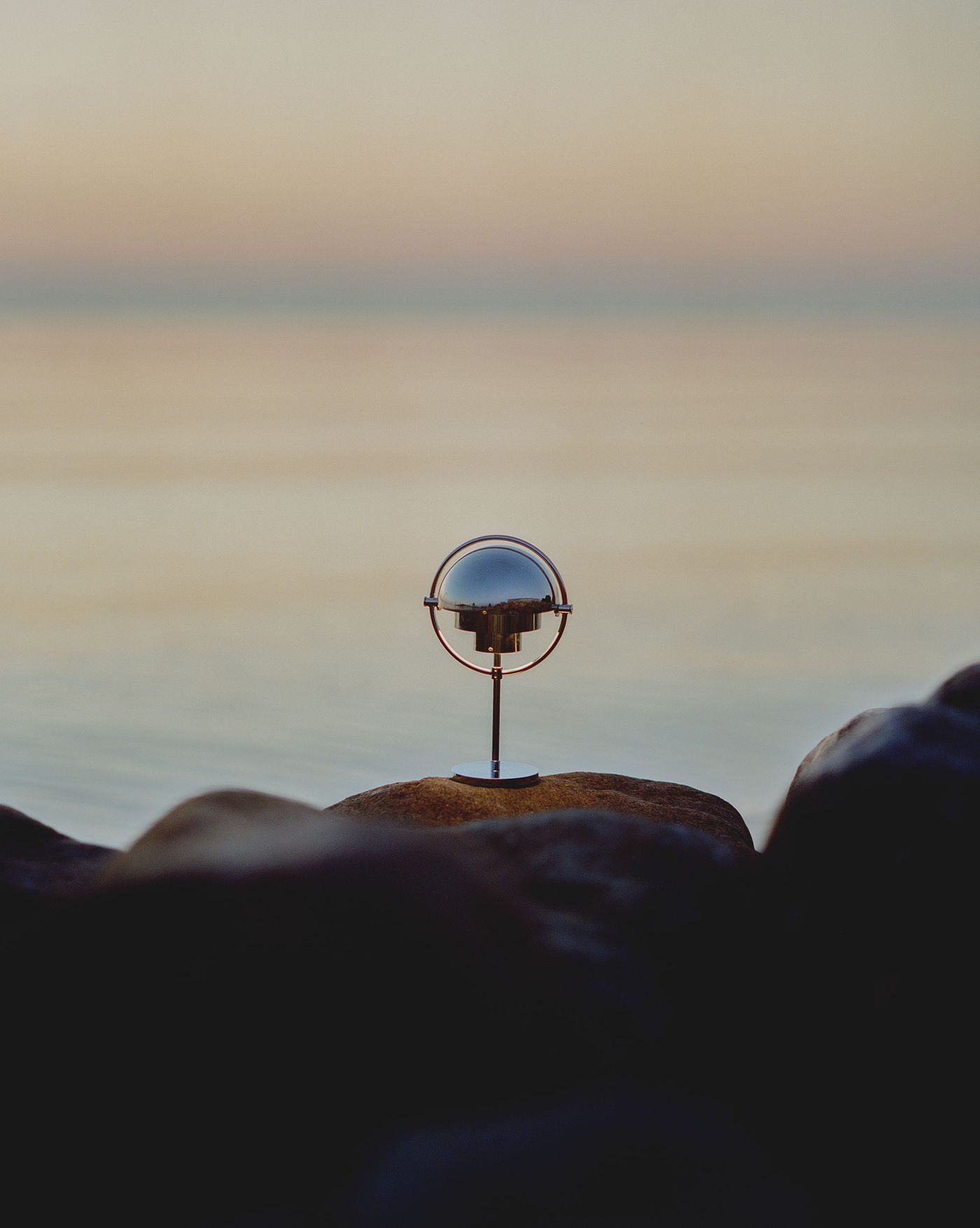 A modern, chrome spherical portable lamp by Gubi with a minimalist design stands on a rock by the edge of calm water at sunset. The lamp's reflective surface mirrors the serene surroundings, and the soft, pastel sky creates a peaceful backdrop. A modern, chrome spherical portable lamp by Gubi with a minimalist design stands on a rock by the edge of calm water at sunset. The lamp's reflective surface mirrors the serene surroundings, and the soft, pastel sky creates a peaceful backdrop.