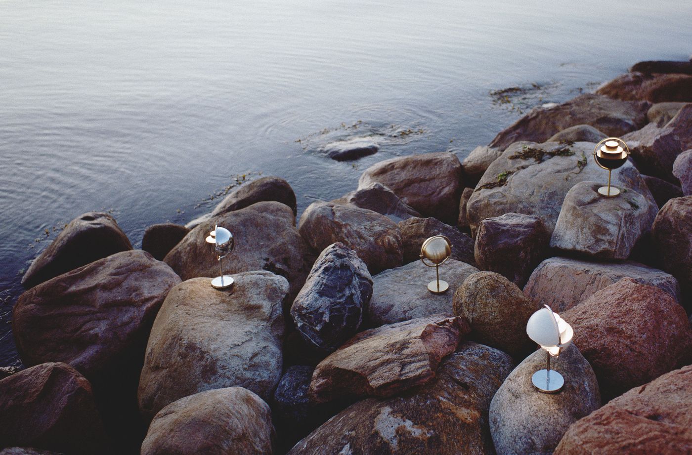 Four modern, spherical portable lamps by Gubi with metallic finishes are placed on large rocks by the edge of a calm body of water. The lamps emit a soft, warm light, contrasting with the natural, rugged texture of the rocks and the serene water in the background. The scene is tranquil and highlights the lamps' contemporary design in an outdoor setting. Four modern, spherical portable lamps by Gubi with metallic finishes are placed on large rocks by the edge of a calm body of water. The lamps emit a soft, warm light, contrasting with the natural, rugged texture of the rocks and the serene water in the background. The scene is tranquil and highlights the lamps' contemporary design in an outdoor setting.