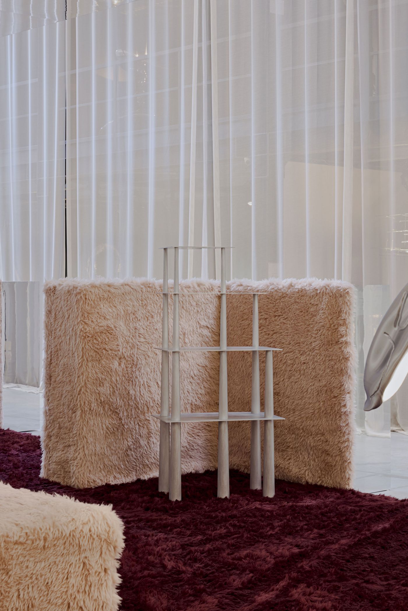 A sculptural, skeletal aluminum shelf stands delicately against plush blush walls and a deep burgundy shag rug—tactile softness meets structural clarity.