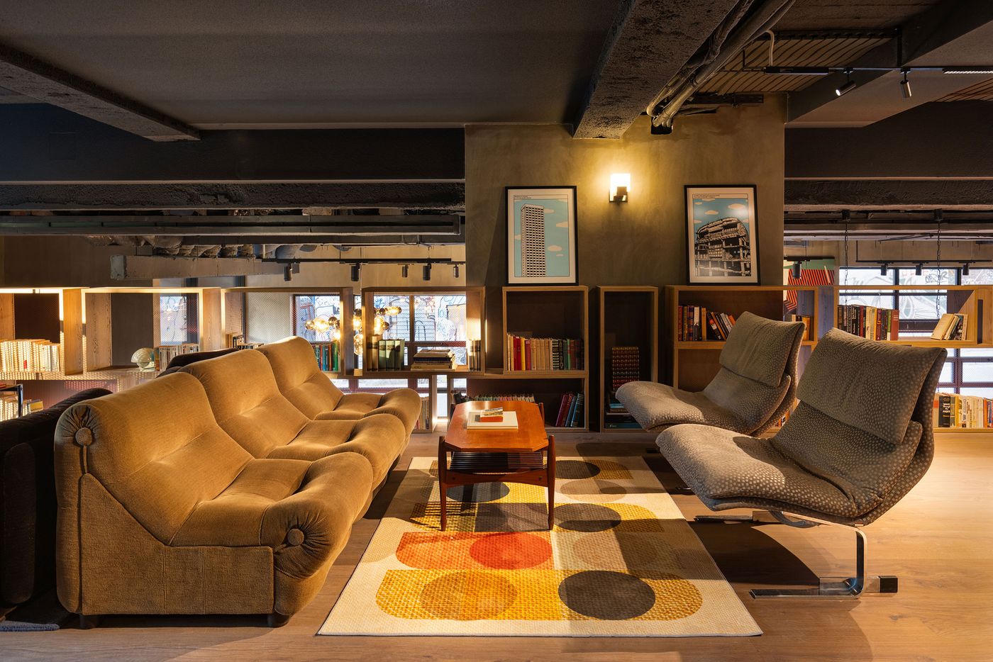 A retro-inspired communal lounge at Okupa hotel in Athens features a mustard velvet sofa, two sculptural upholstered lounge chairs and a vintage timber coffee table arranged over a colourful geometric rug. Open wooden bookshelves, framed architectural prints and warm accent lighting contrast with exposed concrete beams and industrial ceiling details.