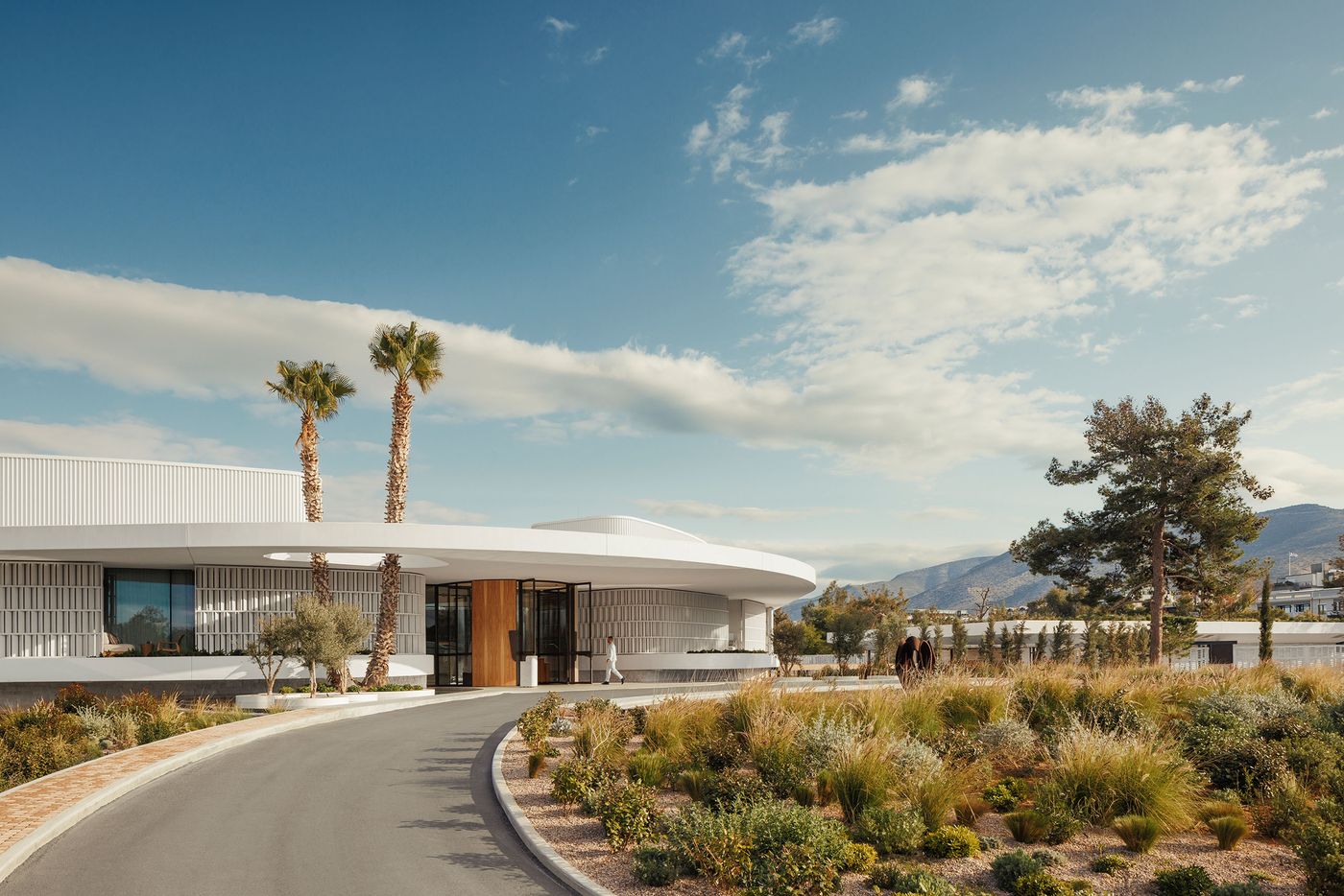 A sweeping view of the modernist architecture at One&Only Aesthesis on the Athens Riviera, featuring a low-slung white building with curved rooflines, vertical façade fins and expansive glazing. A gently curving driveway winds through landscaped Mediterranean gardens, ornamental grasses and palm trees, with pine trees and distant mountains completing the coastal resort setting.