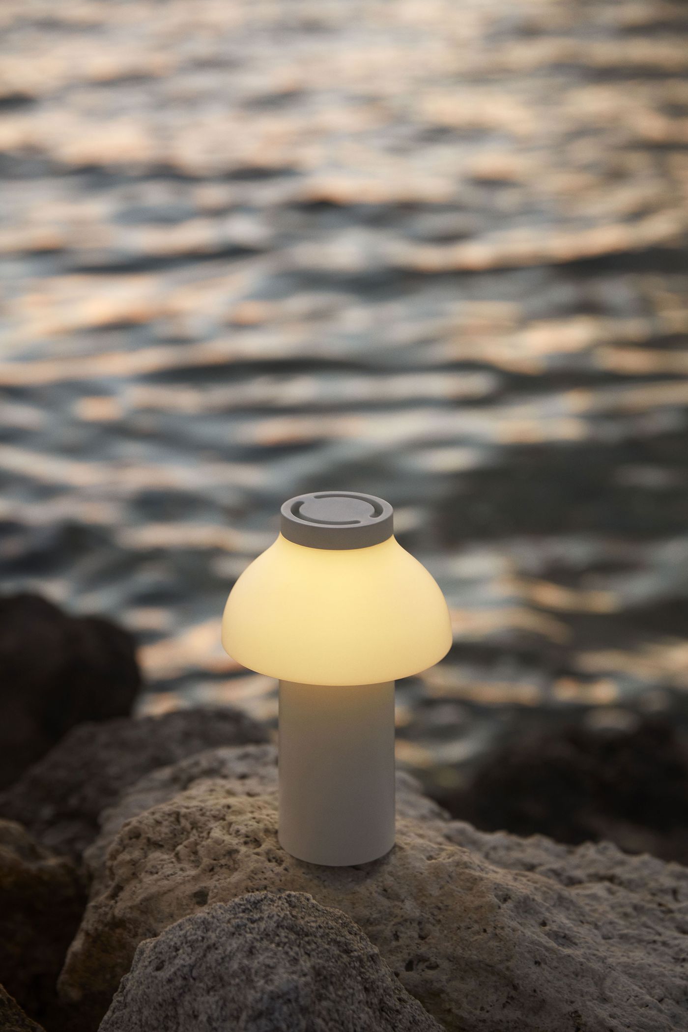 A PC Portable Lamp by HAY softly glows atop a rocky shoreline at dusk, casting warm light against the rippling sea in the background. A PC Portable Lamp by HAY softly glows atop a rocky shoreline at dusk, casting warm light against the rippling sea in the background.