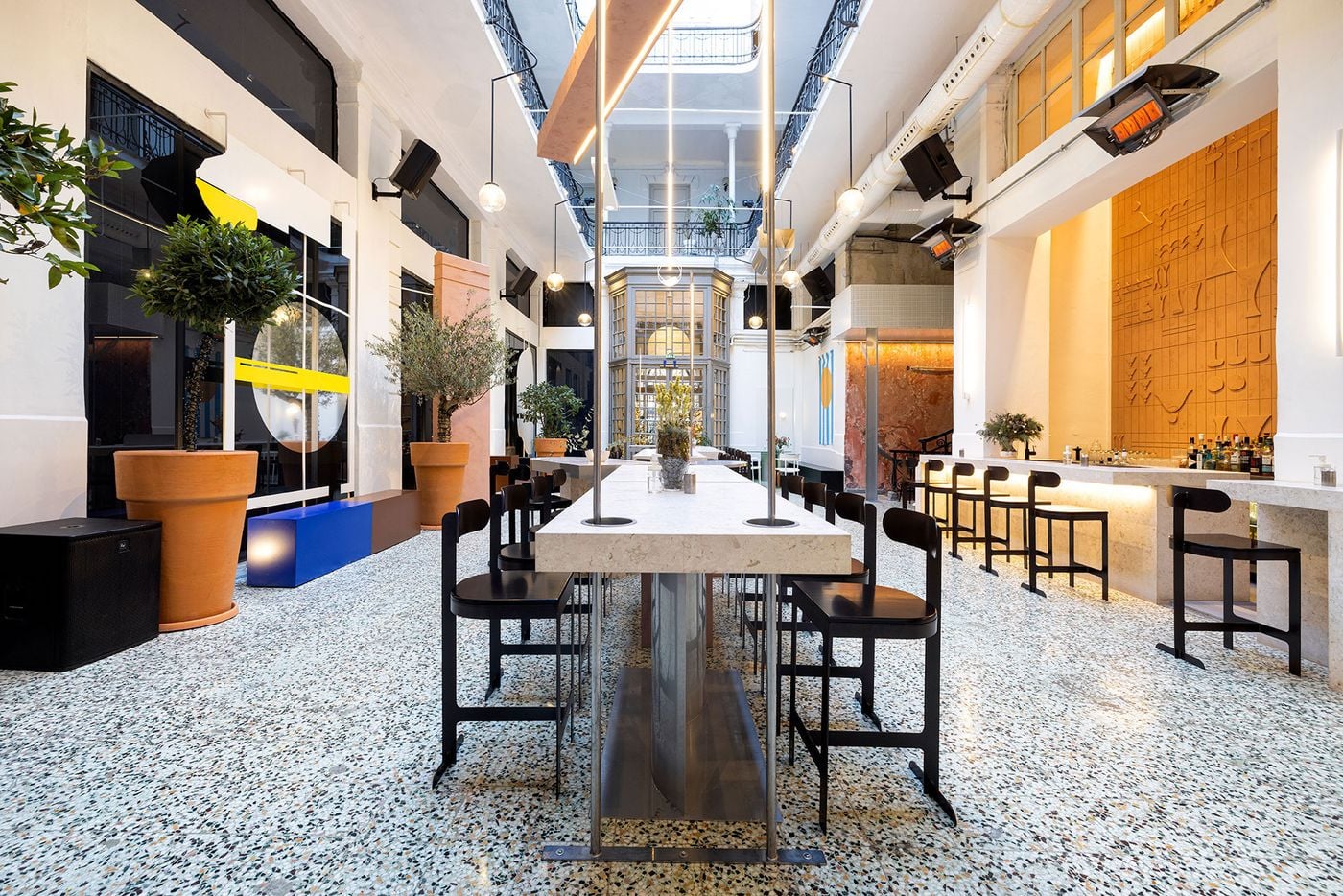 The contemporary interior of Pelosof in Thessaloniki combines a long stone communal table, black wooden chairs, terrazzo flooring and oversized terracotta planters with the historic architecture of a covered arcade. Globe pendant lights, suspended linear fixtures and a striking terracotta relief behind the bar create a layered dialogue between old and new.