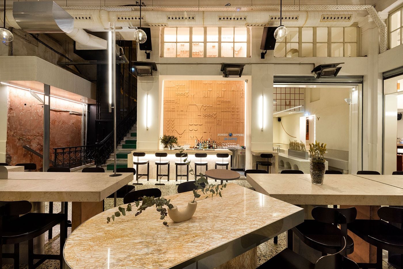 A view of Pelosof’s bar and dining area in Thessaloniki, featuring monumental pale stone tables, black wooden chairs and terrazzo floors. A terracotta relief wall with abstract geometric motifs forms the backdrop to the illuminated bar, while exposed structural elements, suspended globe lights and a historic staircase add architectural depth.