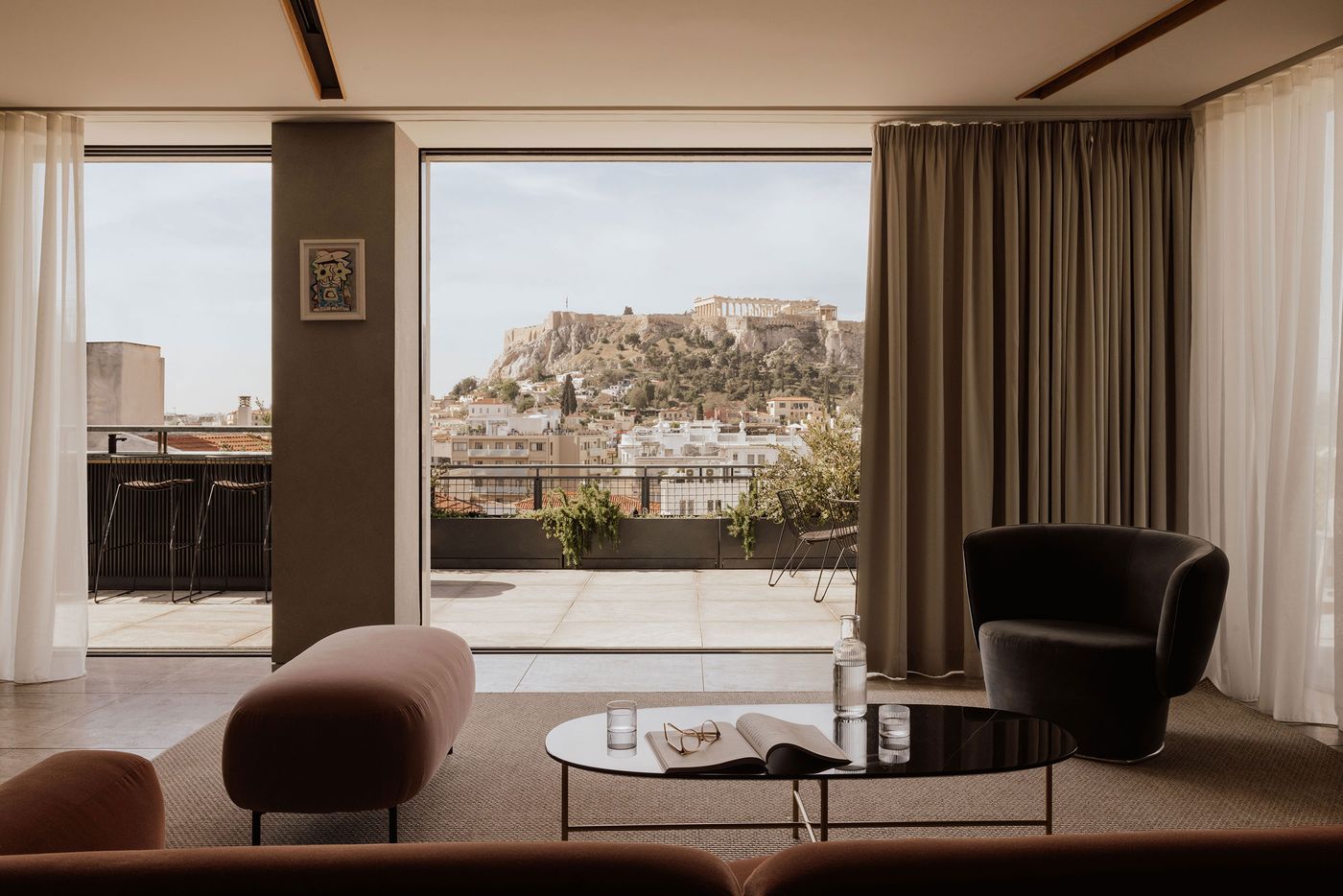 A contemporary suite at Perianth Hotel in Athens opens onto a spacious private terrace overlooking the Acropolis and Parthenon. Floor-to-ceiling windows frame the historic skyline, while a sculptural black armchair, blush-toned upholstered bench and low oval coffee table create a restrained, modernist interior in muted earthy tones.