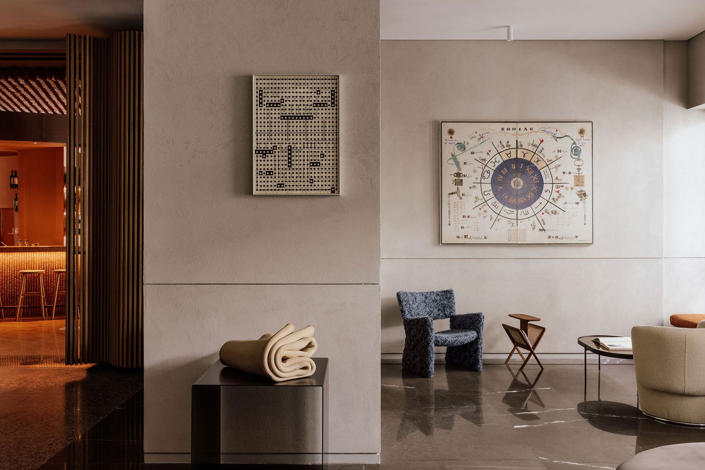 An art-filled lounge at Perianth Hotel in Athens combines polished dark marble floors, pale textured walls and eclectic contemporary furnishings. A tactile blue armchair, sculptural folded ceramic piece and geometric timber side table are complemented by graphic artworks, while a glimpse of the warmly illuminated bar introduces a contrasting amber glow.