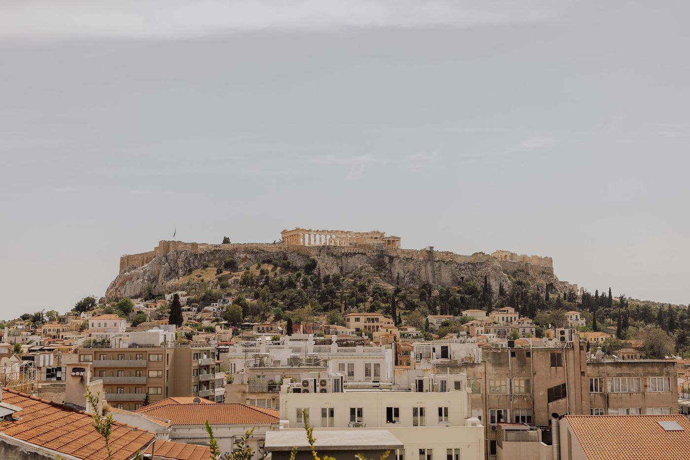 A panoramic view of the Acropolis and Parthenon rising above the densely layered rooftops of central Athens, photographed from Perianth Hotel. Classical ruins and ancient stone fortifications crown the rocky hill, contrasting with the surrounding mix of neoclassical houses, terracotta roofs and modern apartment buildings beneath a softly overcast sky.