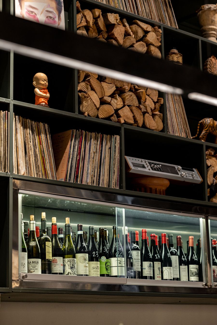 A close-up of the eclectic shelving at Pharaoh restaurant in Athens reveals an assemblage of vinyl records, stacked firewood, vintage audio equipment and playful decorative objects, including a small monk figurine and a striped tiger. A glass-fronted wine cabinet beneath the dark metal shelving reinforces the restaurant's distinctive combination of analogue music culture and rustic character.