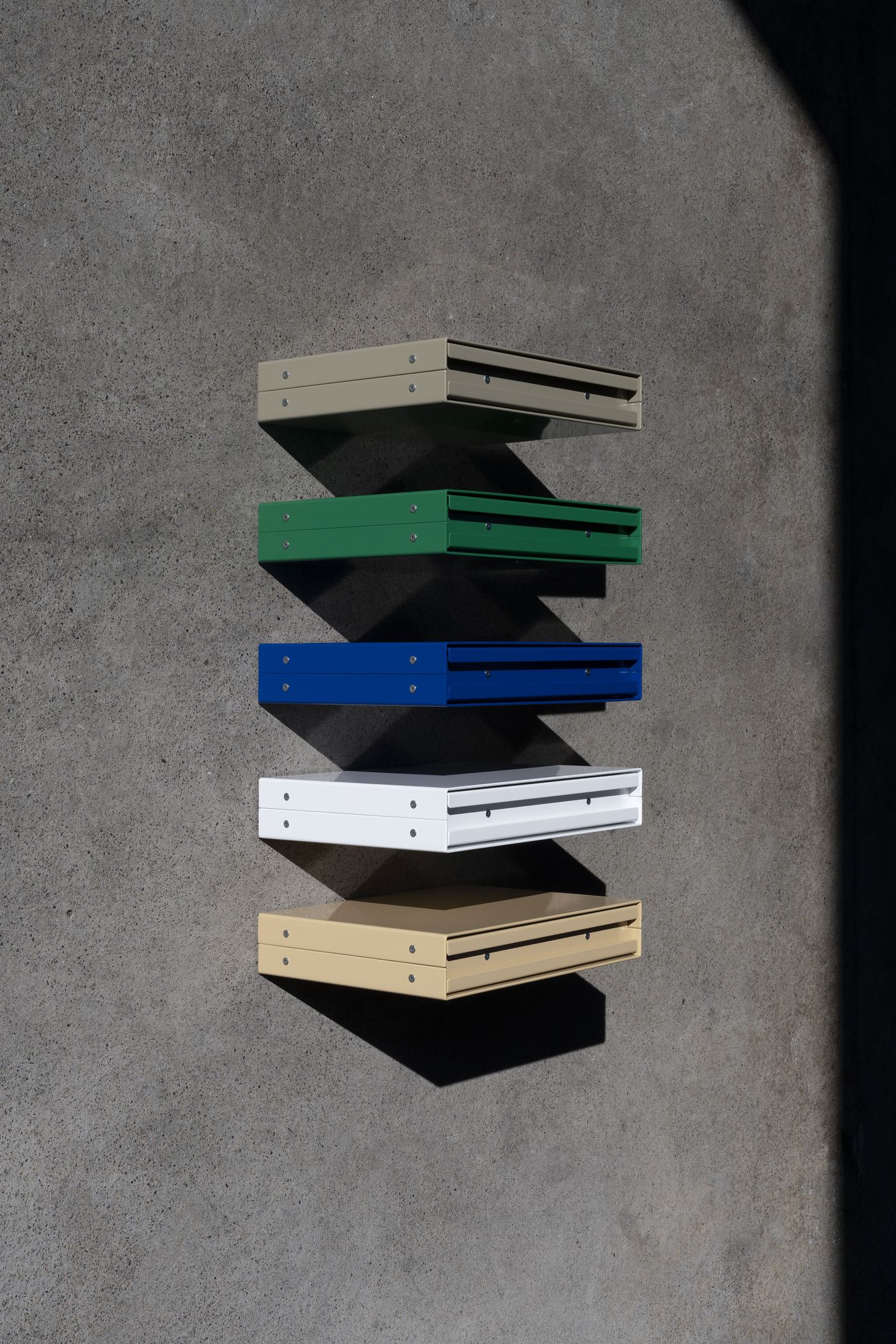 Five wall-mounted metal storage units in beige, green, blue, white, and pale yellow, shown in sharp light and shadow against a raw concrete wall—minimalist geometry meets industrial color play. Five wall-mounted metal storage units in beige, green, blue, white, and pale yellow, shown in sharp light and shadow against a raw concrete wall—minimalist geometry meets industrial color play.