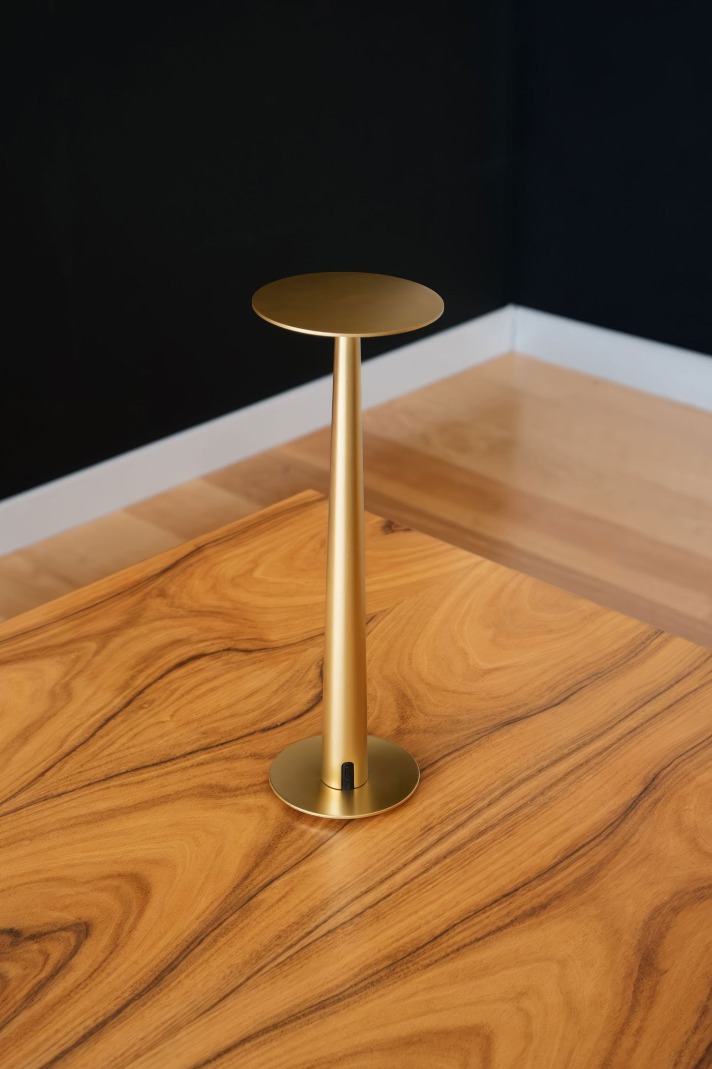 A sleek, gold-colored, minimalist table lamp with a slender conical stem and round top, placed on a wooden table with a swirling grain pattern, set against a black wall and light wood floor.
