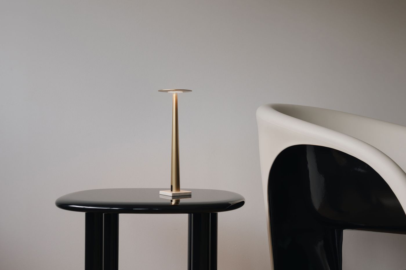 A modern, gold-colored table lamp with a slim, tapered stem and circular top, standing on a glossy black side table next to a sculptural white and black chair, with a plain light gray wall in the background.