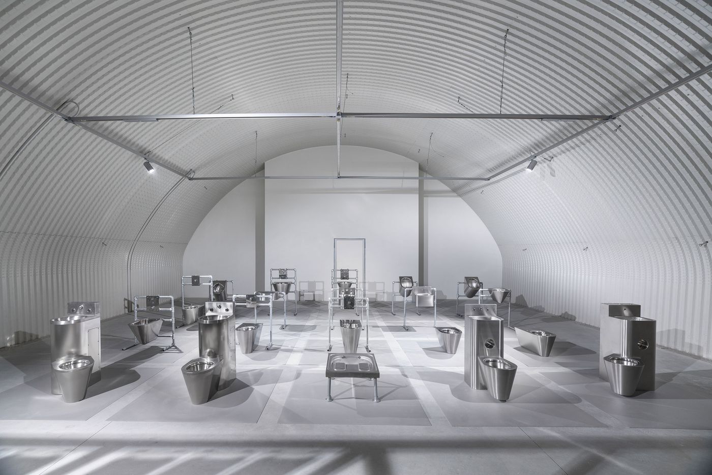 Industrial-style exhibition of stainless steel prison toilets and sinks arranged in a symmetrical grid under a corrugated vault.