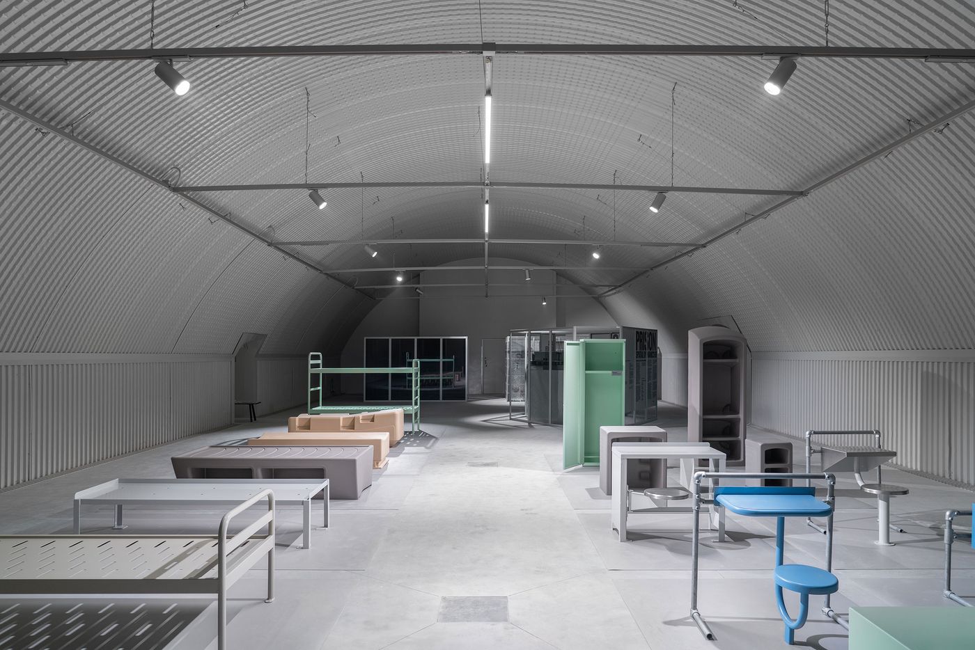 Display of pastel-colored prison furniture, including beds, tables, and lockers, in a minimalist arched warehouse.