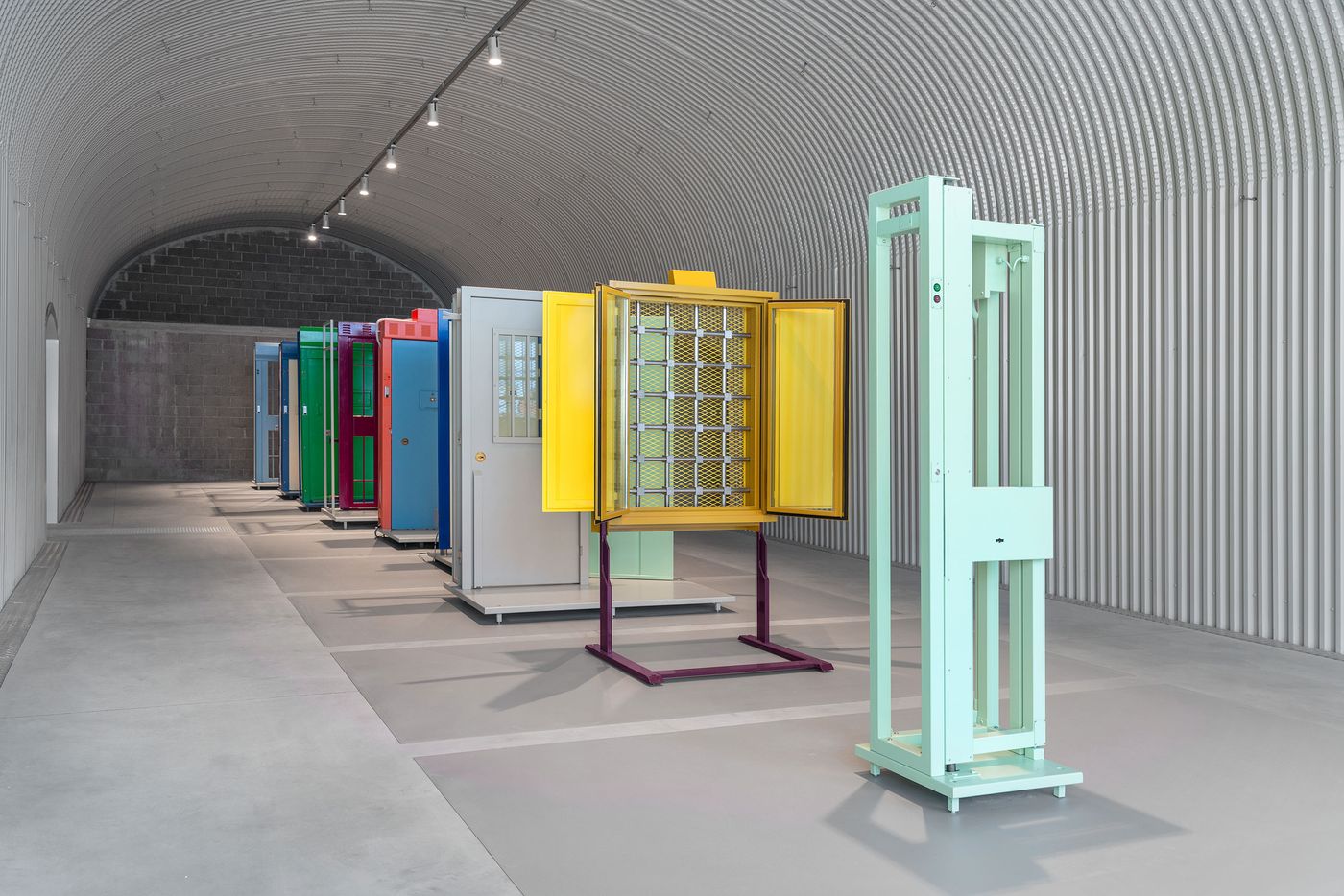 Prison cafeteria setup with mint-green and blue tables facing a vibrant wall of multicolored plastic food trays and utensils.