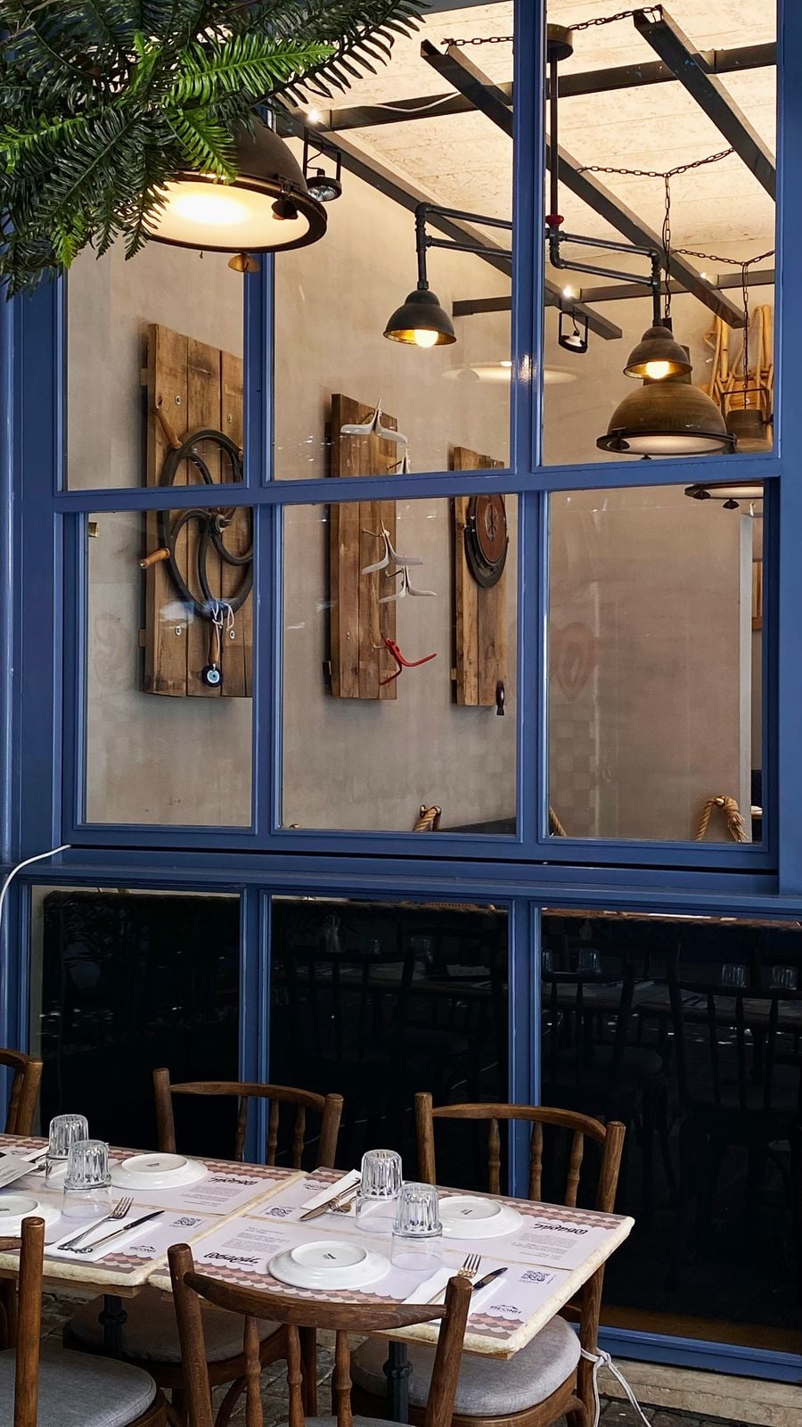 A glimpse of Psaras restaurant in Thessaloniki through its blue-painted window frames reveals a rustic interior with nautical-inspired decorations mounted on timber panels, exposed steel beams and industrial pendant lights. Outside, wooden chairs and neatly laid tables create an inviting, traditional taverna atmosphere.