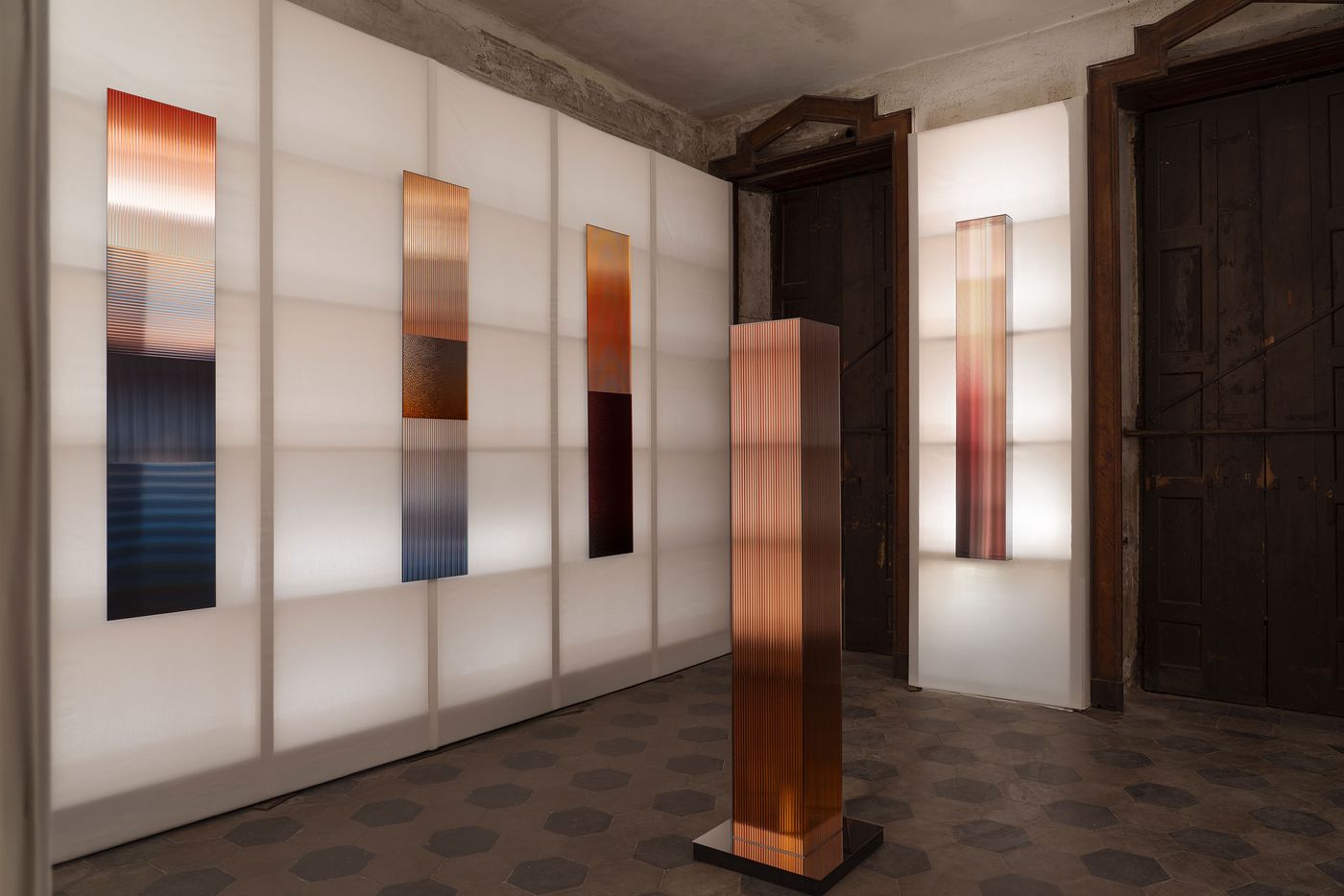 A moody interior featuring tall, rectangular glass sculptures with gradient colors, mounted on softly lit white fabric walls and a freestanding matching sculpture in the center.