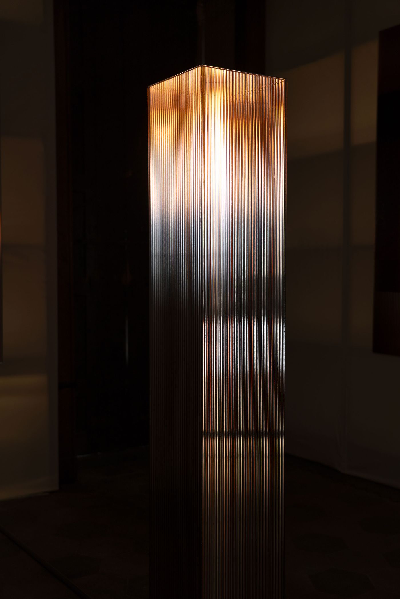 A glowing vertical glass column with fine ribbed texture, radiating warm light in a dimly lit, rustic space.