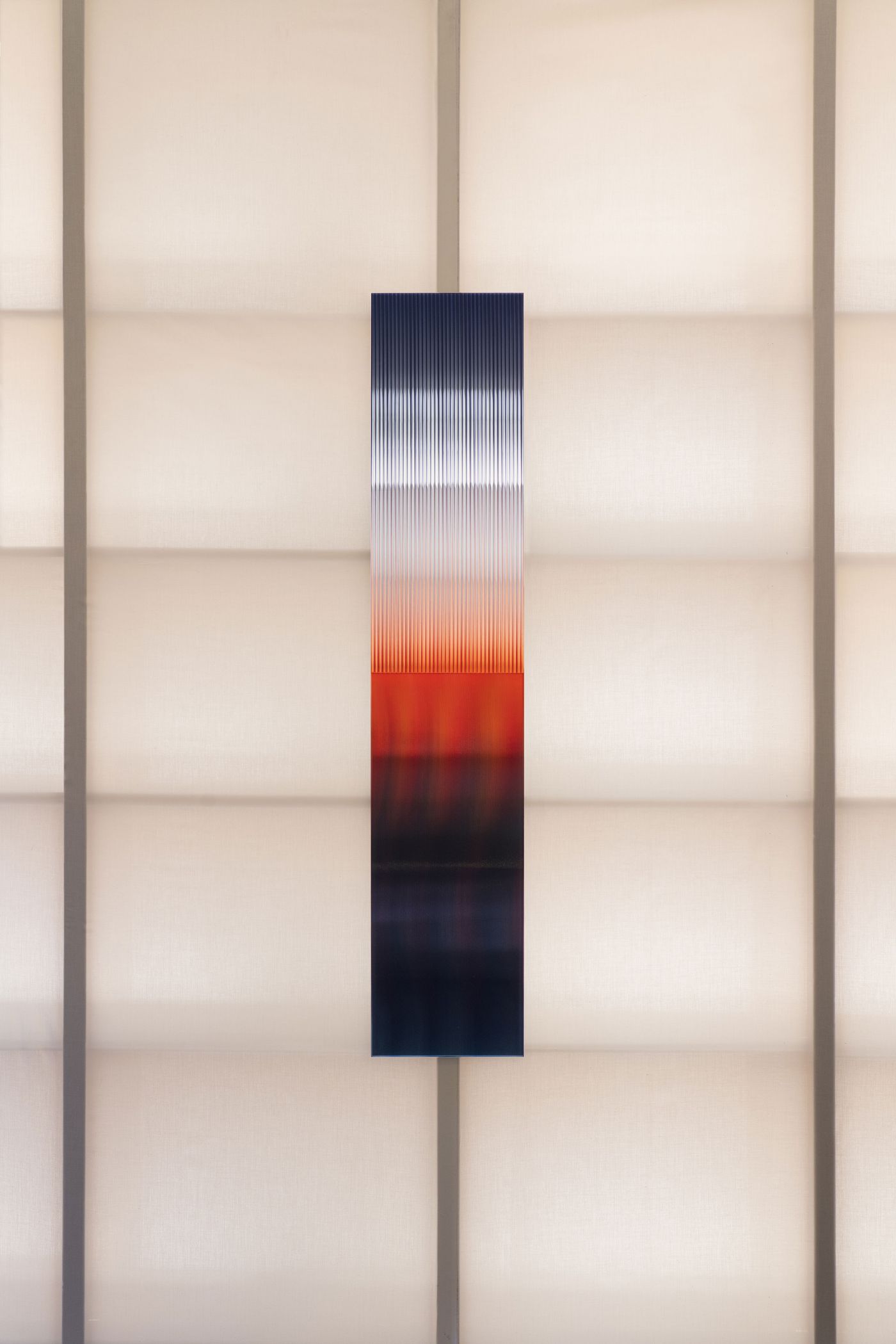 A singular vertical gradient glass panel blending blue, white, and red hues, mounted on a grid-like white fabric wall.