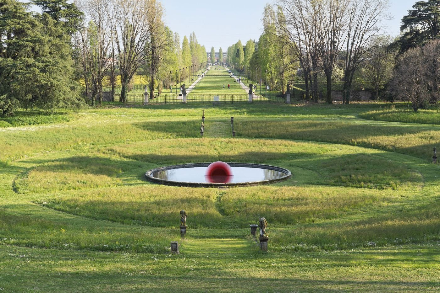 A striking landscape view of a historic Italian garden with manicured paths, leading lines of trees, and a central round pond holding a glowing red hemisphere sculpture. A striking landscape view of a historic Italian garden with manicured paths, leading lines of trees, and a central round pond holding a glowing red hemisphere sculpture.