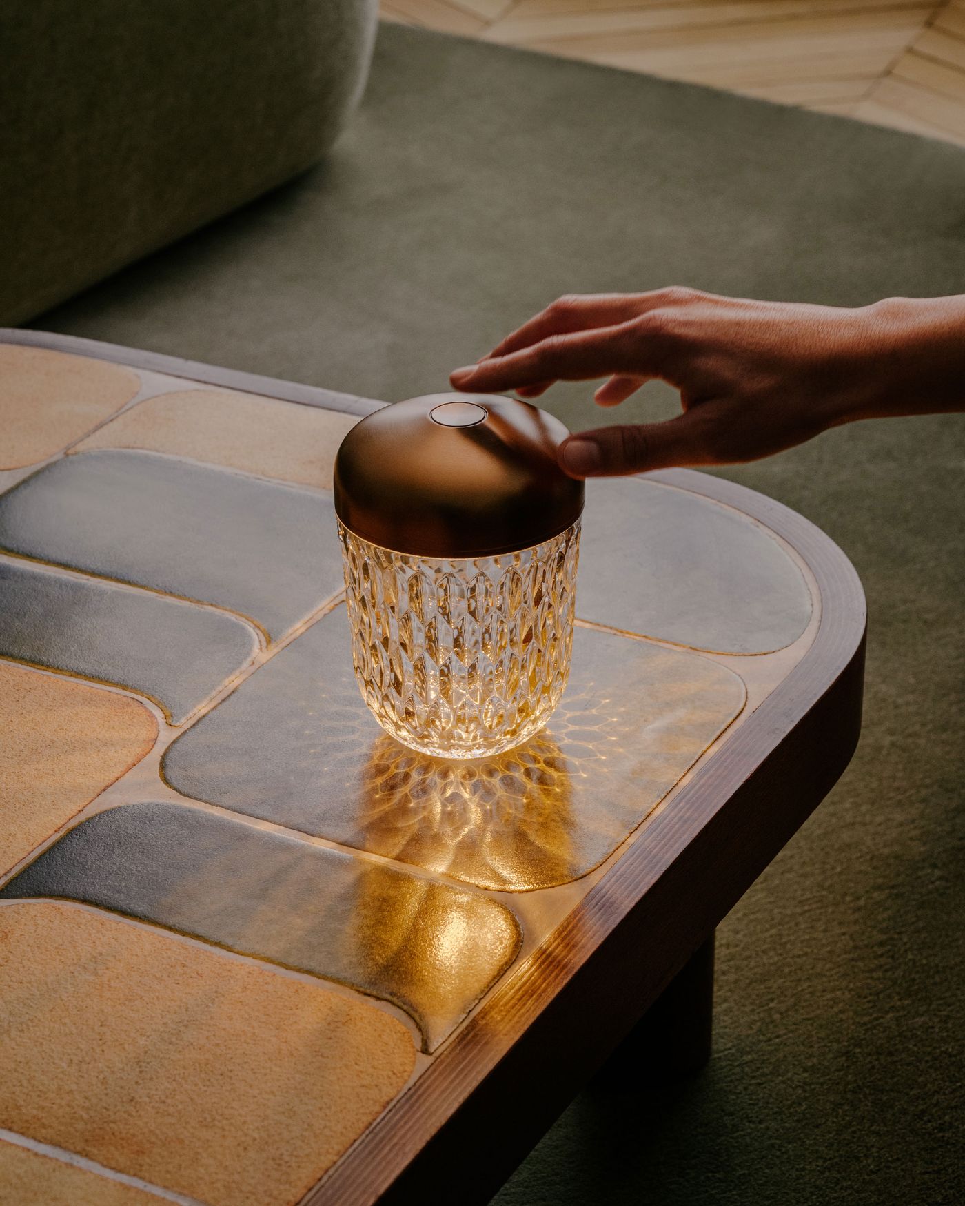A hand reaches to touch a crystal table lamp with a bronze top, casting intricate light reflections on a glazed tile coffee table.