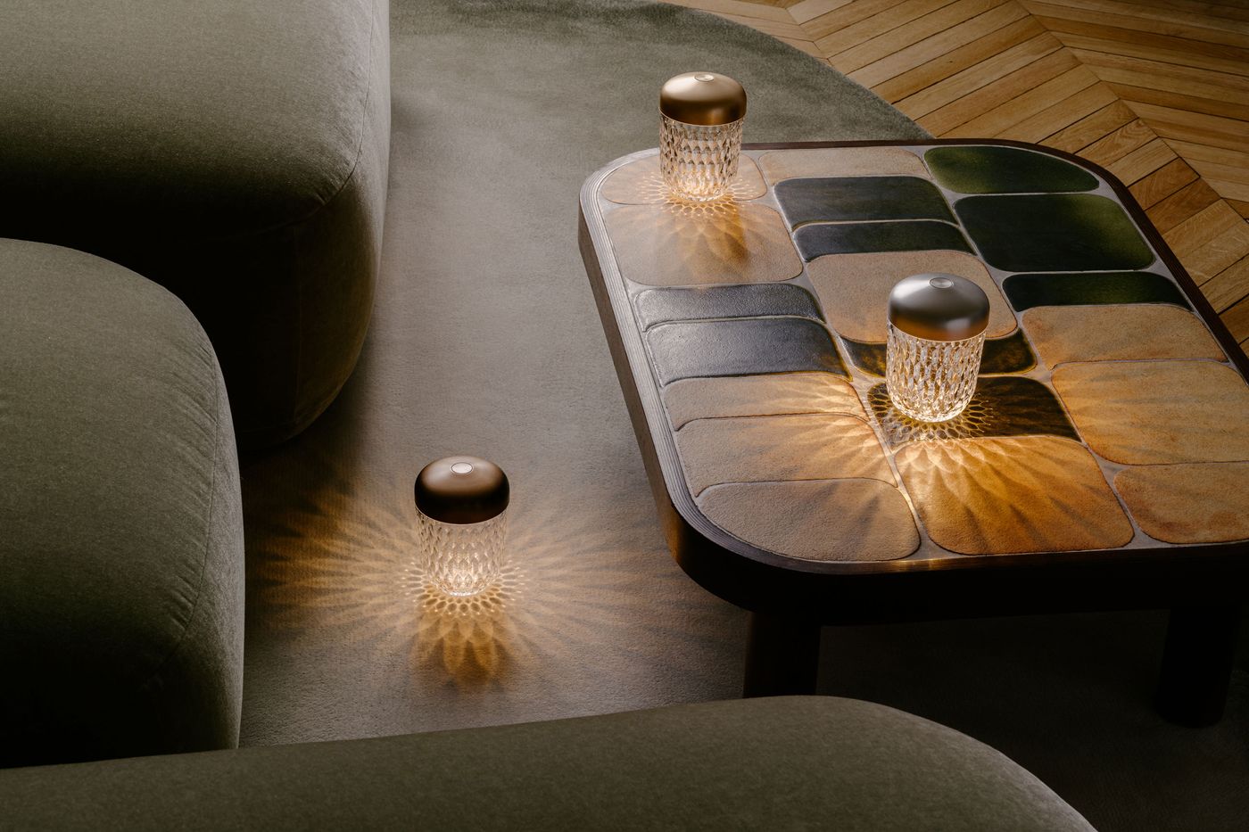 Three crystal table lamps with metallic tops glow warmly on and around a tiled coffee table, surrounded by soft green seating.