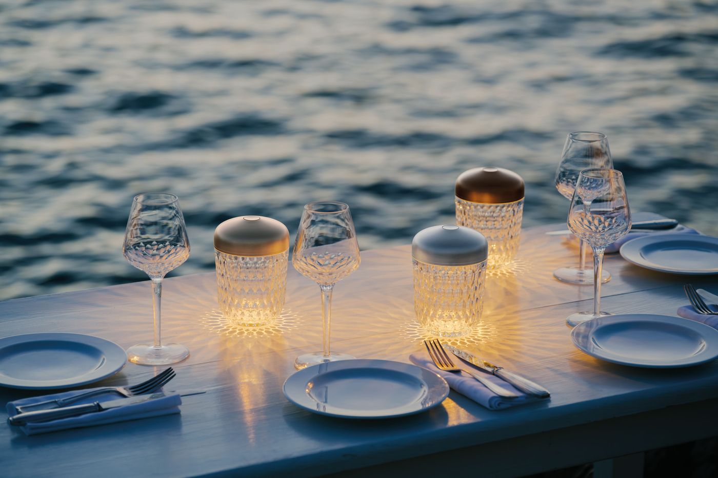 A set of crystal lamps with bronze and silver tops illuminate a seaside dining table at dusk, reflecting patterns through wine glasses and plates. A set of crystal lamps with bronze and silver tops illuminate a seaside dining table at dusk, reflecting patterns through wine glasses and plates.