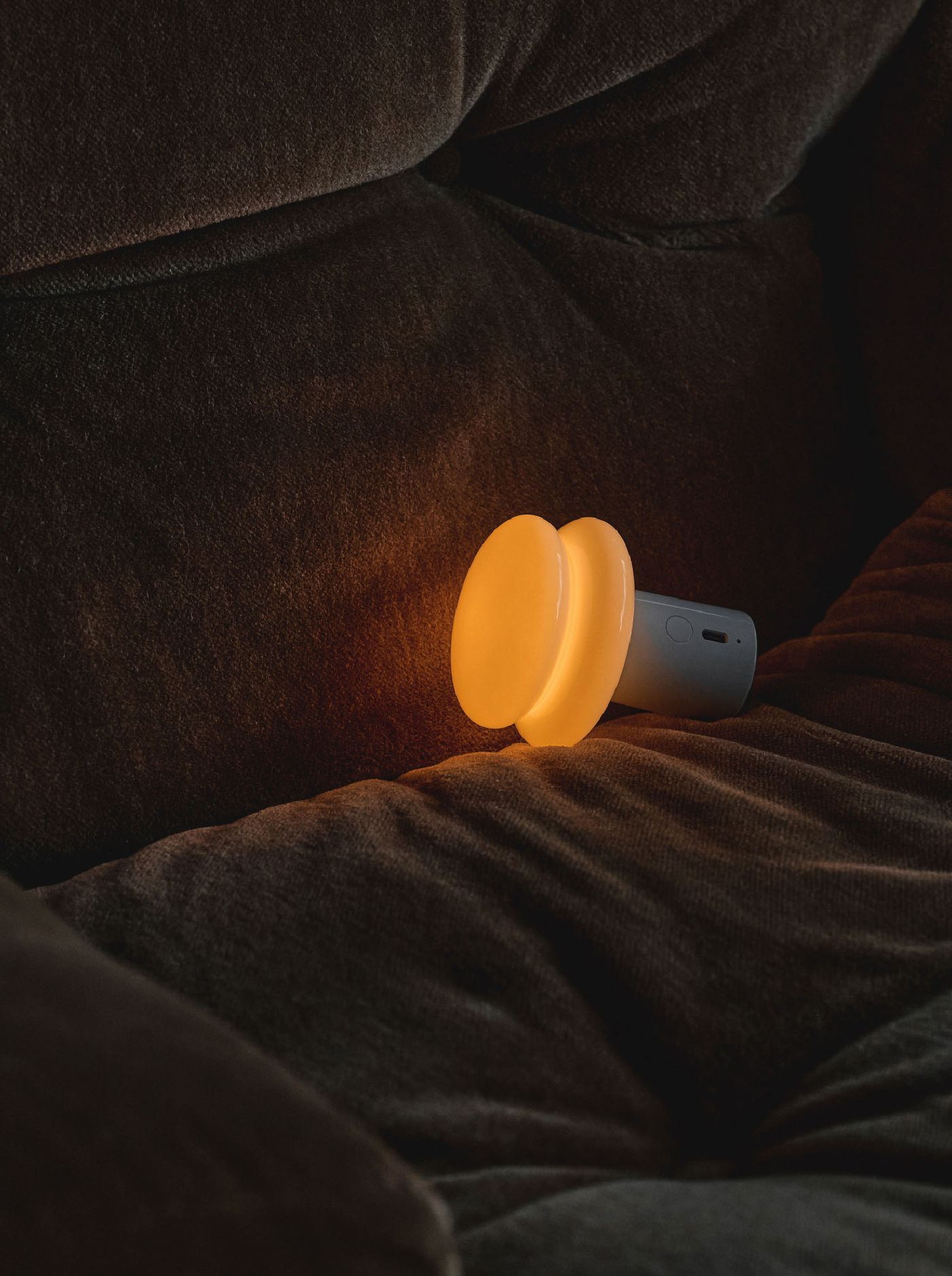 The Snowman8 lamp emits a cozy amber light as it rests on a deep brown velvety couch, blending charm with comfort.