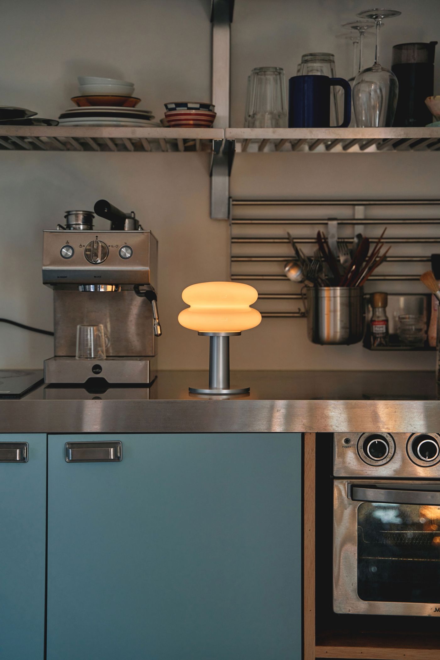 A softly glowing table lamp with a double-layered rounded glass form from the SNOWMAN series by ilkwdesign, placed on a kitchen counter next to a coffee machine, creating a cozy ambient light.