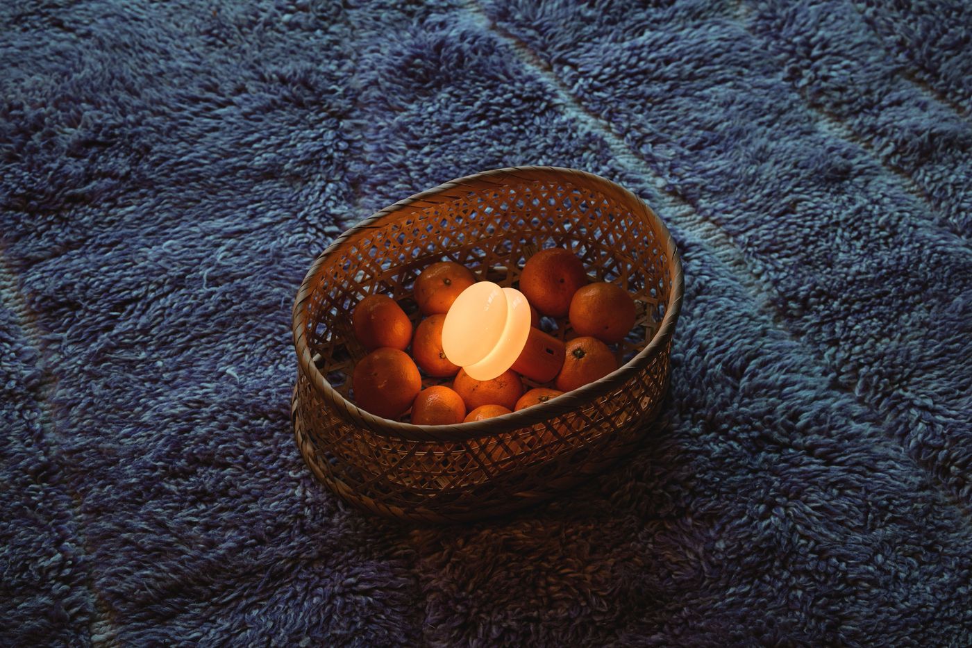 A small glowing SNOWMAN lamp nestled among mandarins in a woven basket on a textured blue rug, blending playful form with functional lighting.