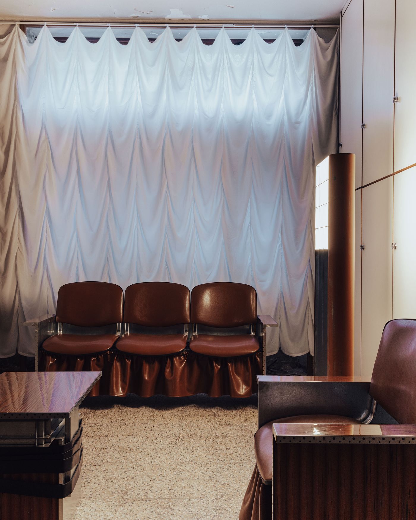 A retro-futuristic lounge featuring a trio of brown leather and metal chairs with gathered skirts, a glossy wood and steel table, and a floor lamp, all set against dramatic, ruched white curtains.